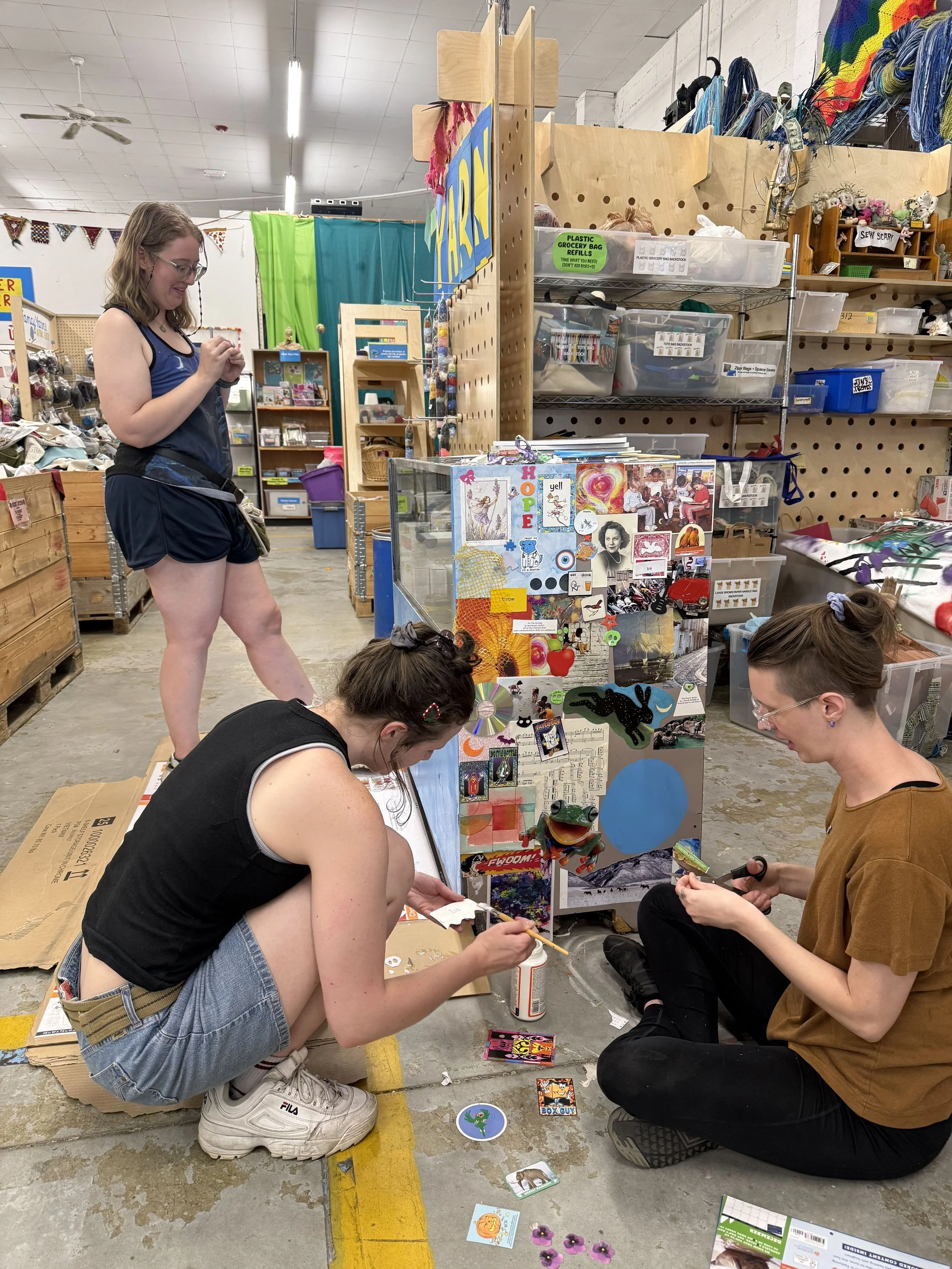 Three women participating in an arts and crafts activity in a warehouse-like space. Two are sitting on the floor working on craft projects, while one stands nearby. The space has shelves with various craft supplies, colorful decorations, and scattered craft items on the floor.