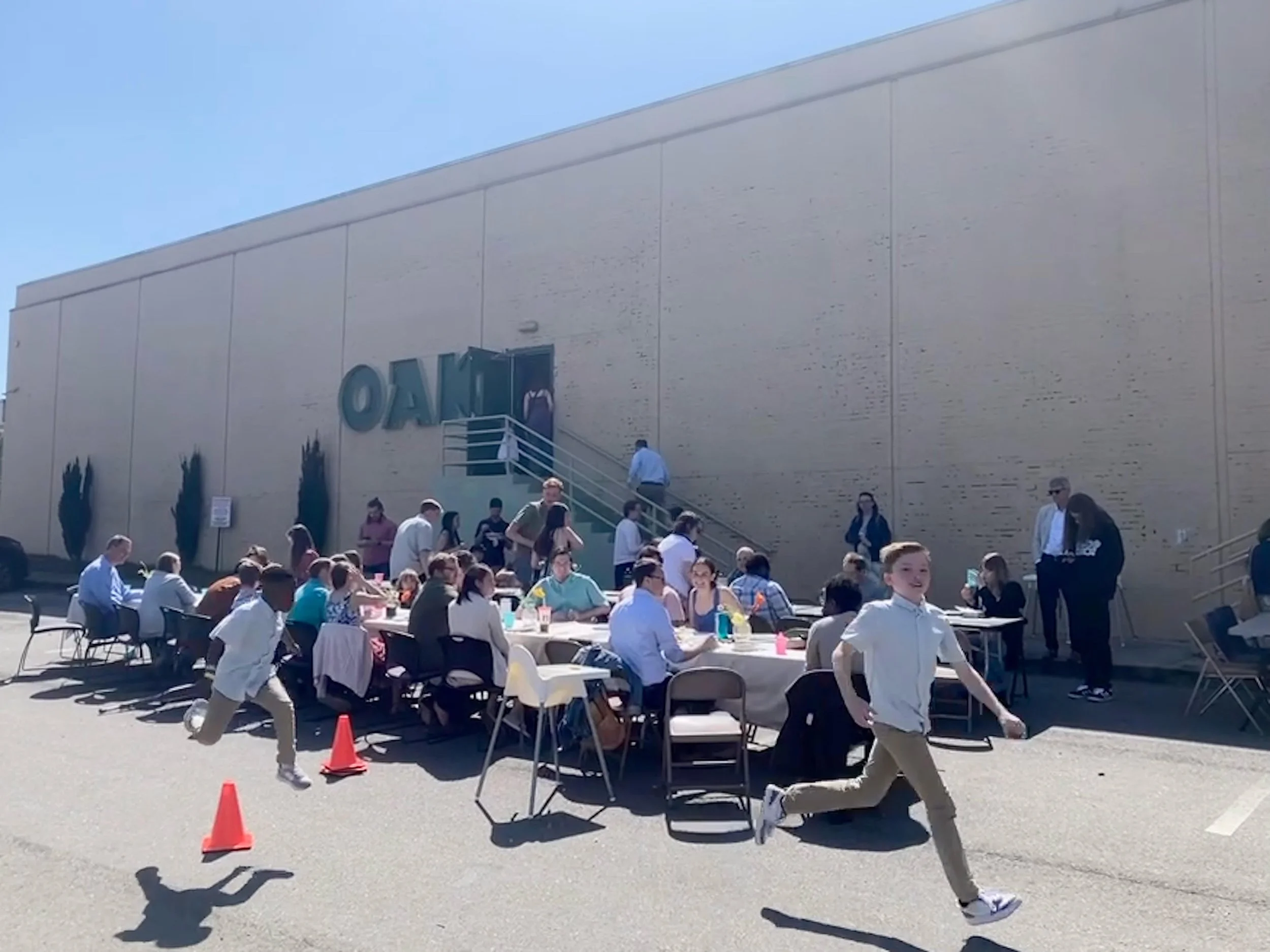 People are gathered outdoors around tables with chairs, participating in an event near a large building with a beige wall. Some individuals are walking, while others are seated or standing, enjoying the sunny day.