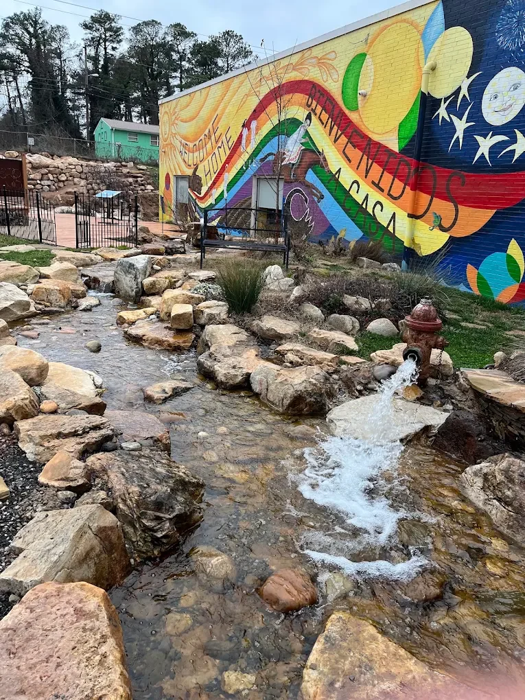Small creek with rocks and flowing water in front of a colorful mural with a rainbow, sun, and stars, and a building with a bench and trees in the background.