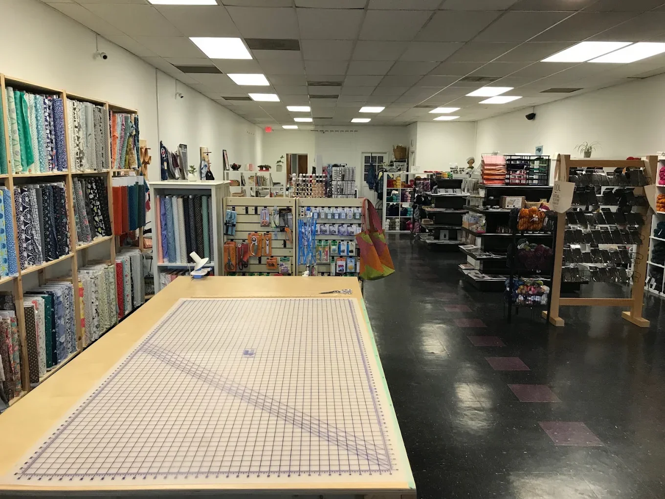 Interior of a craft or fabric store with shelves of fabric bolts on the left, and various craft supplies and yarn on the right. A large cutting mat is on the table in the foreground.