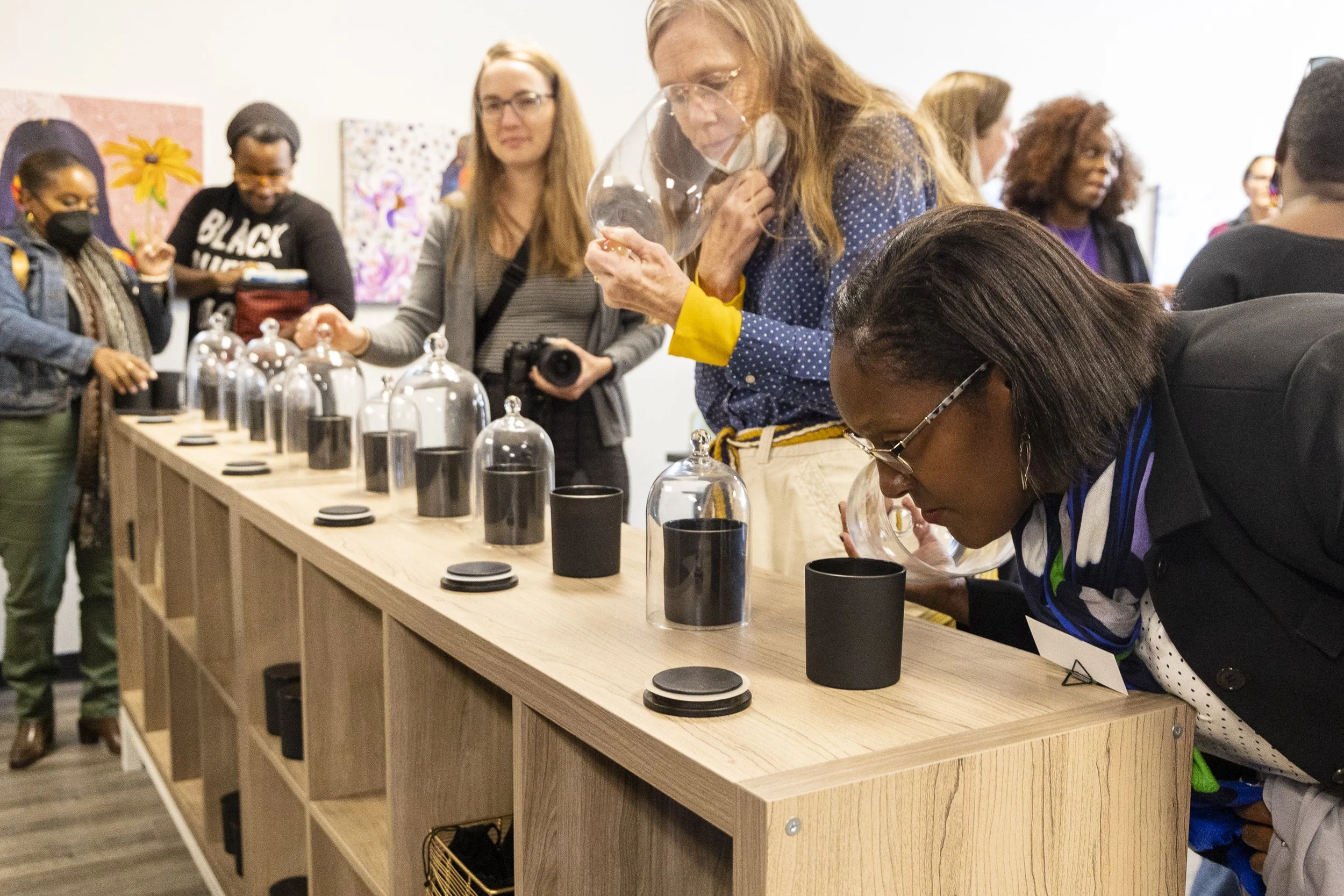 Women participating in a scent testing or fragrance workshop, smelling glass containers on a wooden table, with artworks on the wall behind them.
