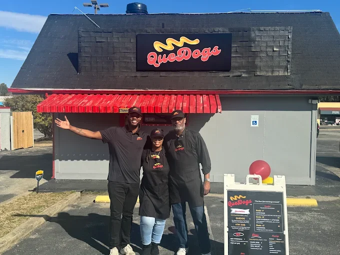 Three people standing outside a QueDogs restaurant, smiling, with one person extending their arm. The restaurant has a black roof, a red awning, and a sign with a yellow hot dog illustration and red text. There is a menu board on the sidewalk.
