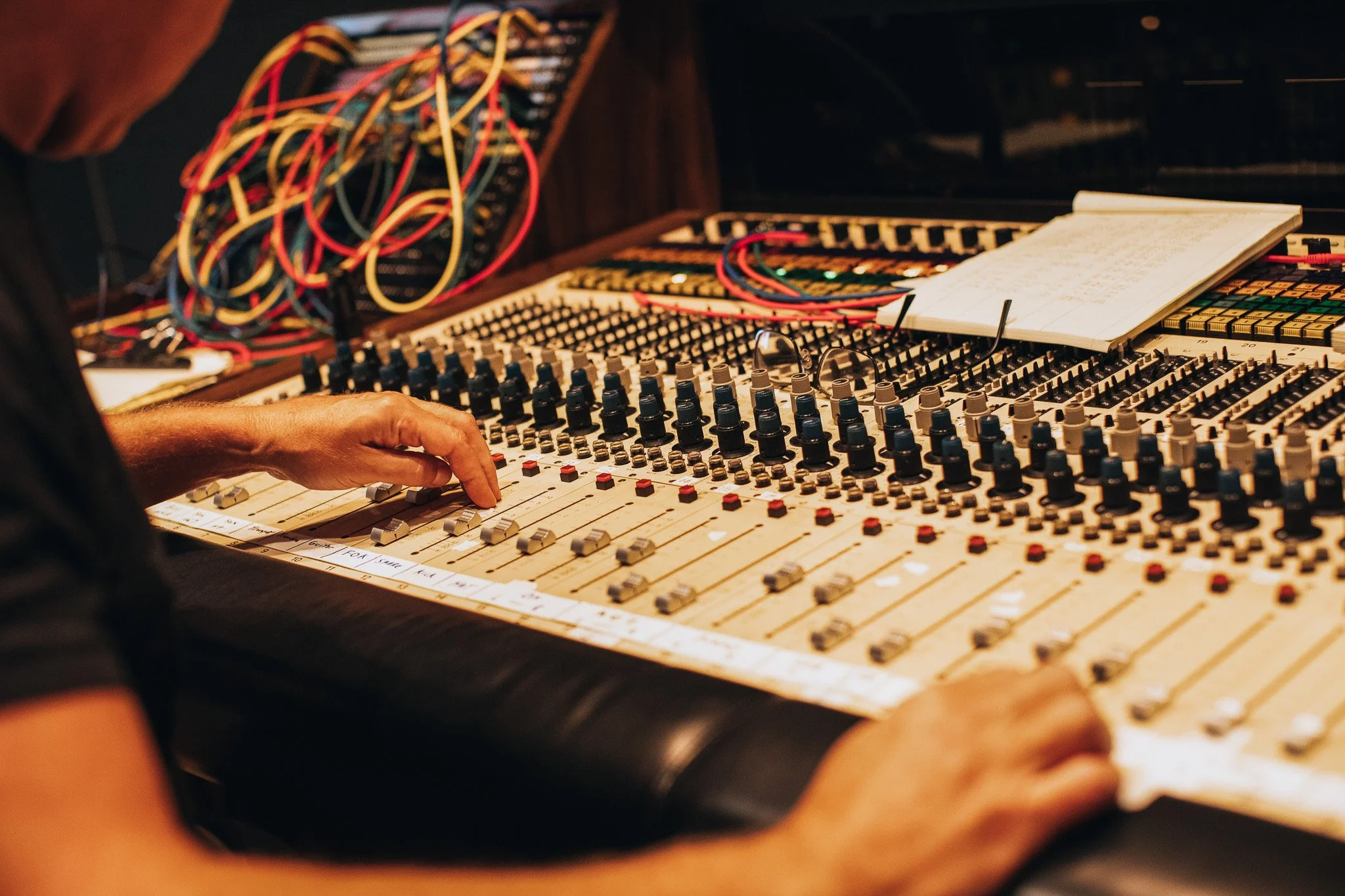 A person's hand adjusting controls on a large audio mixing console with numerous knobs, sliders, and patch cables, in a recording studio or sound engineering setting.