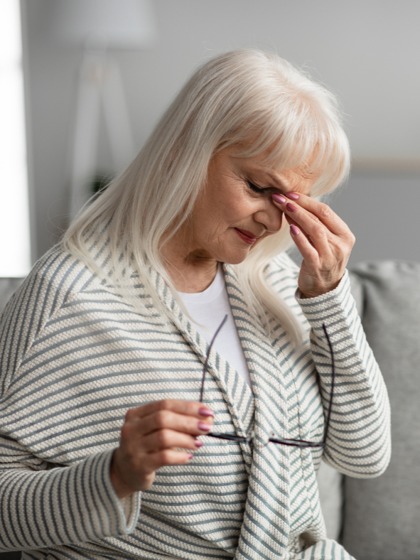 Older woman sitting on a couch holding her glasses and appearing stressed, representing the emotional toll caregiving and aging concerns can place on families.