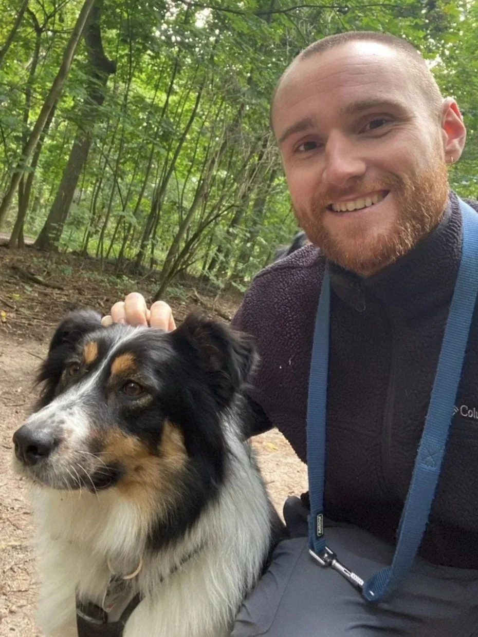 A smiling man with a beard and a blue lead, wearing outdoor clothing, taking a selfie with an Australian Shepherd dog in a wooded area.