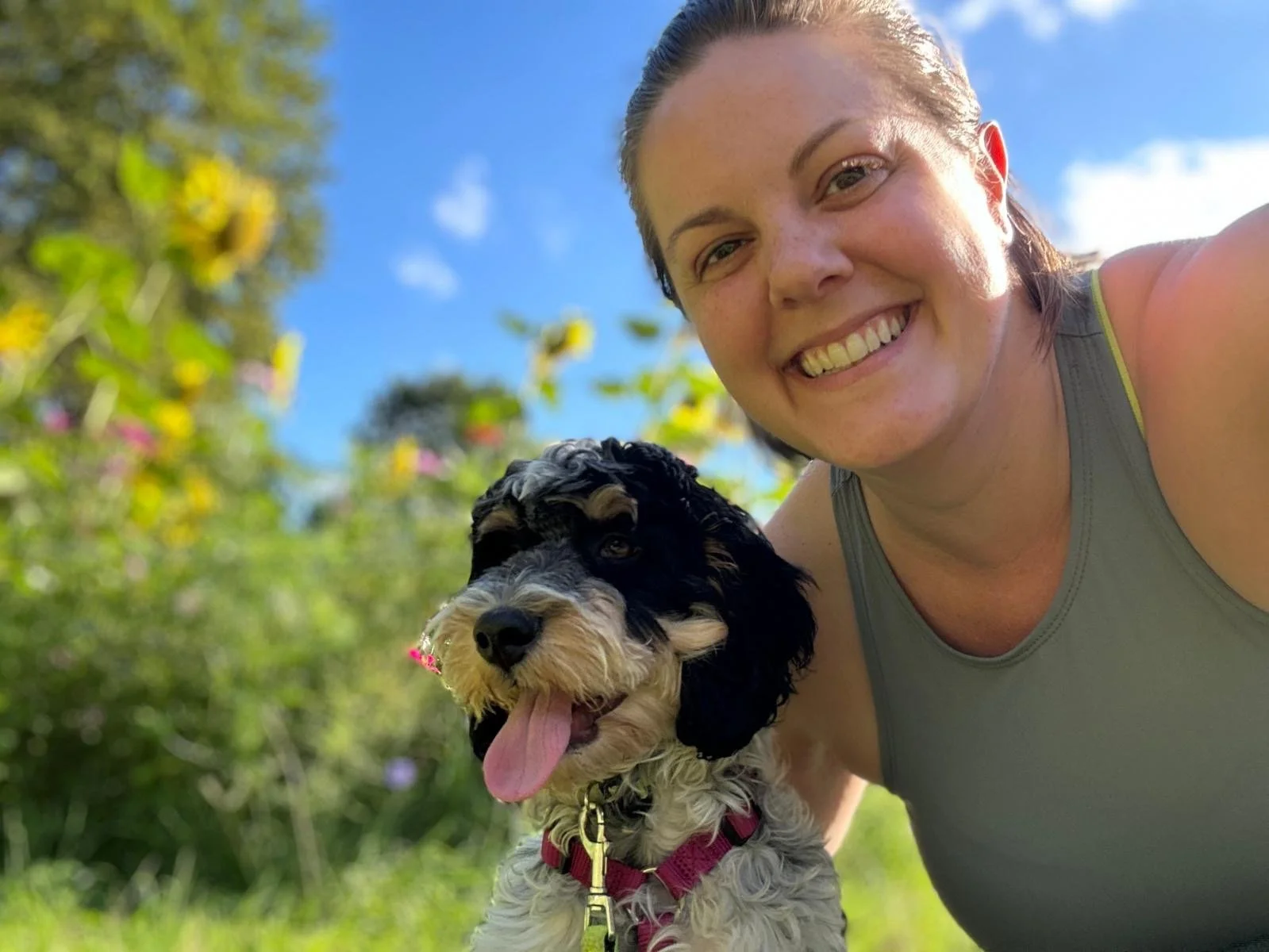 A woman smiling while taking a selfie with her dog outdoors on a sunny day, with trees and blue sky in the background.