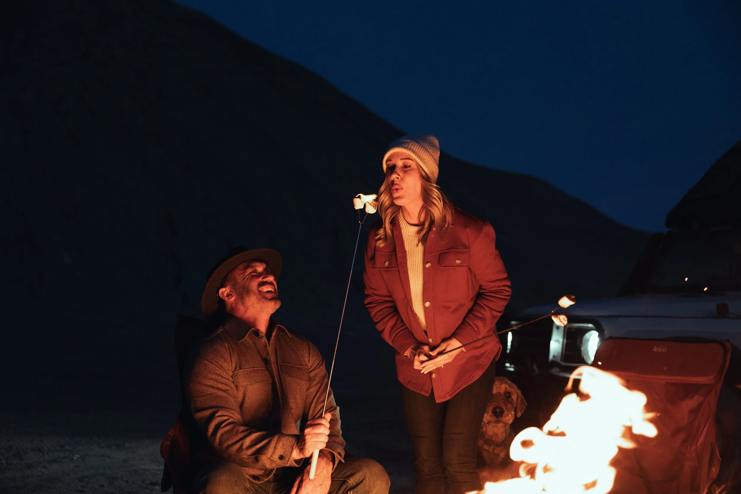 A man and a woman camping at night around a campfire. The man, wearing a brown jacket and hat, is sitting and laughing, while the woman, wearing a red jacket and a knit beanie, is standing and blowing a marshmallow on a stick. There is a dog behind t