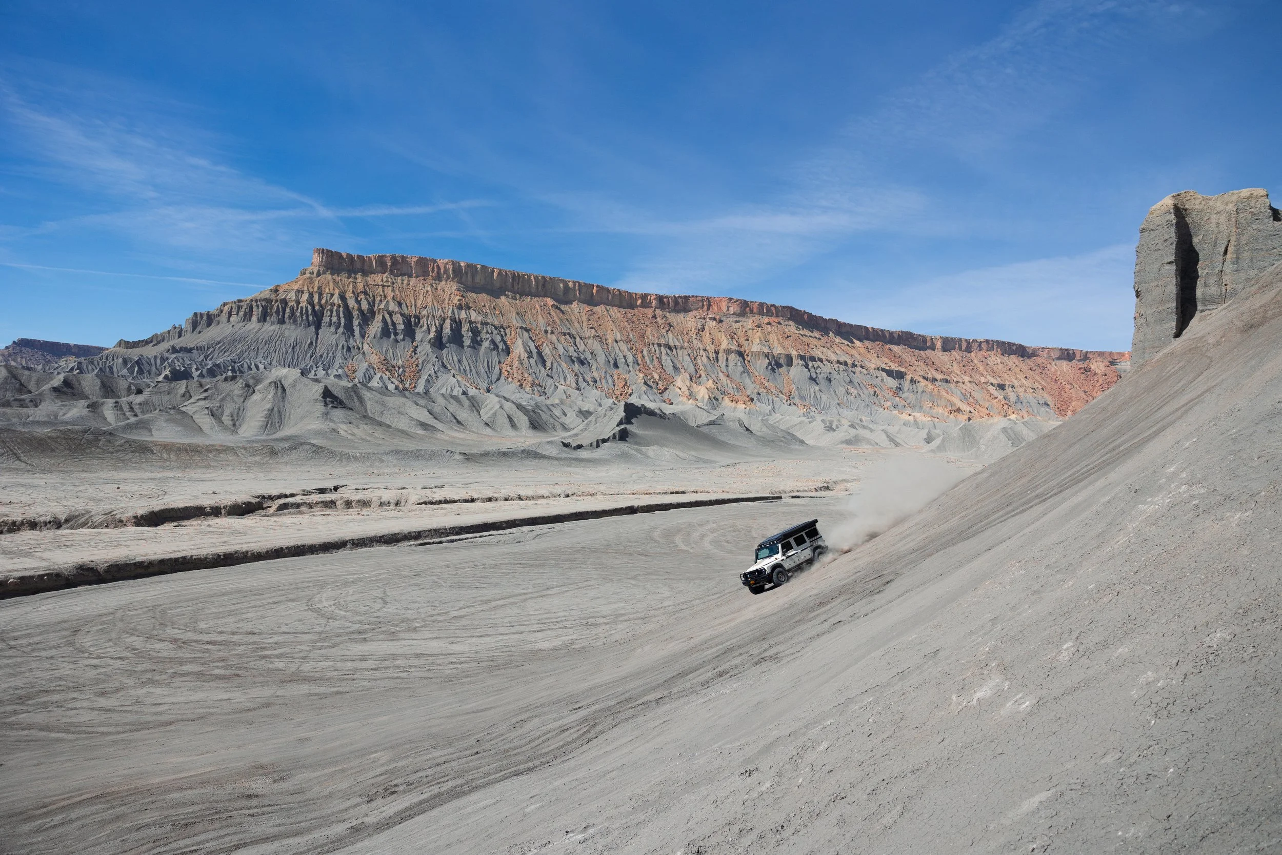 A vehicle driving on a steep, dusty slope in a desert landscape near a canyon with layered rock formations, under a clear blue sky.