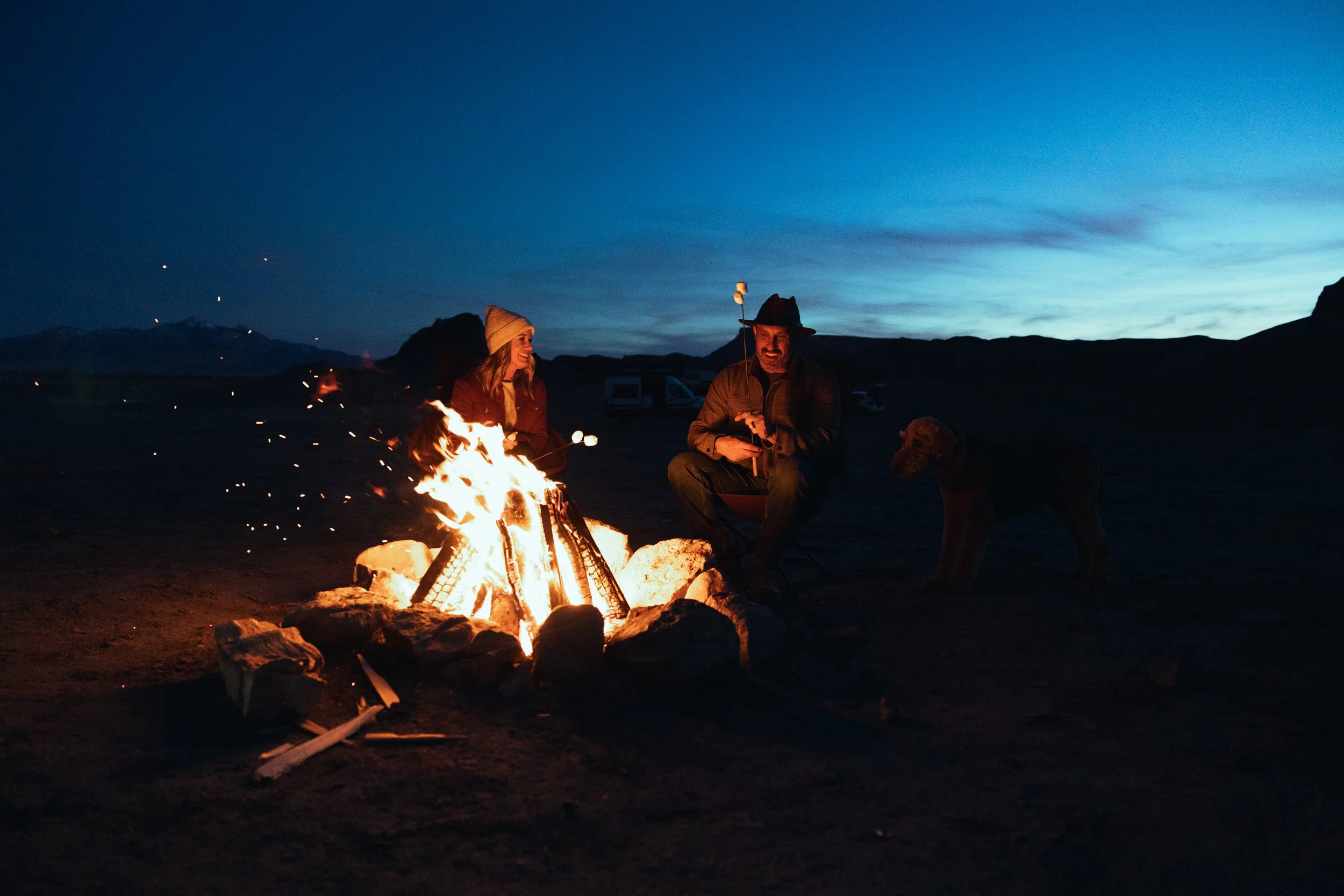 Two people and a dog sitting around a campfire at dusk in a mountain landscape.