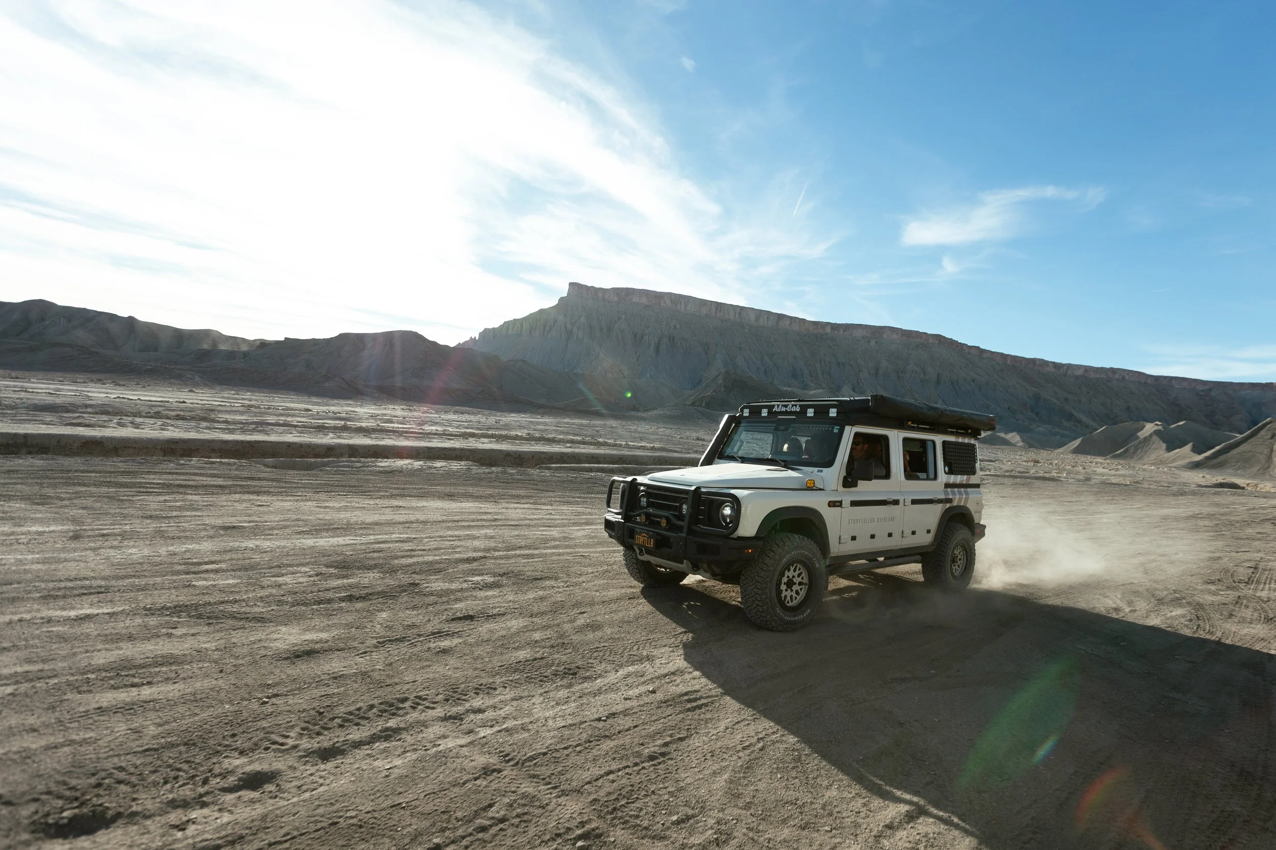 A white off-road vehicle driving on a dusty desert landscape with mountains and a blue sky in the background.