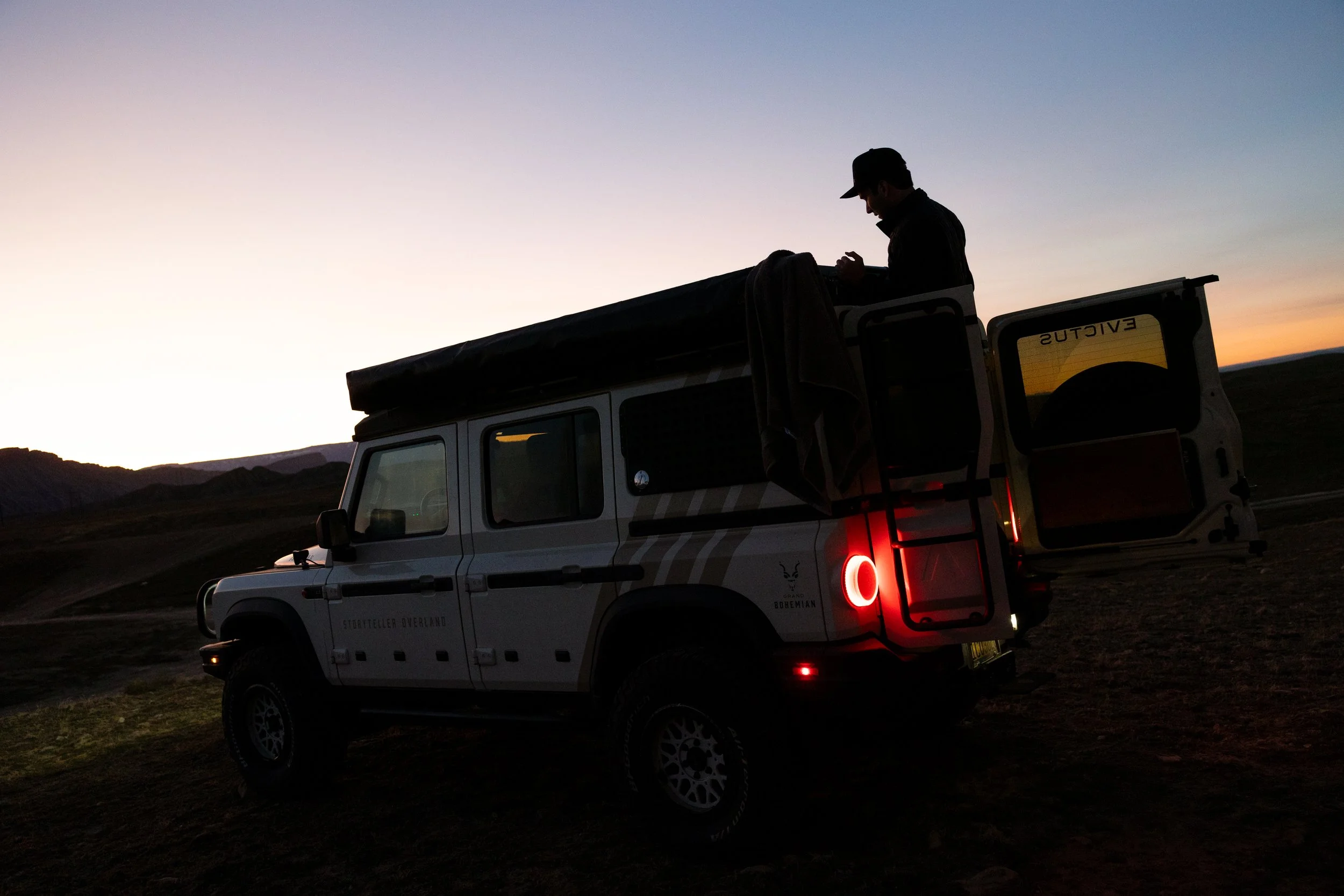 A person standing on the back of an off-road vehicle during sunset in a remote, mountainous landscape.