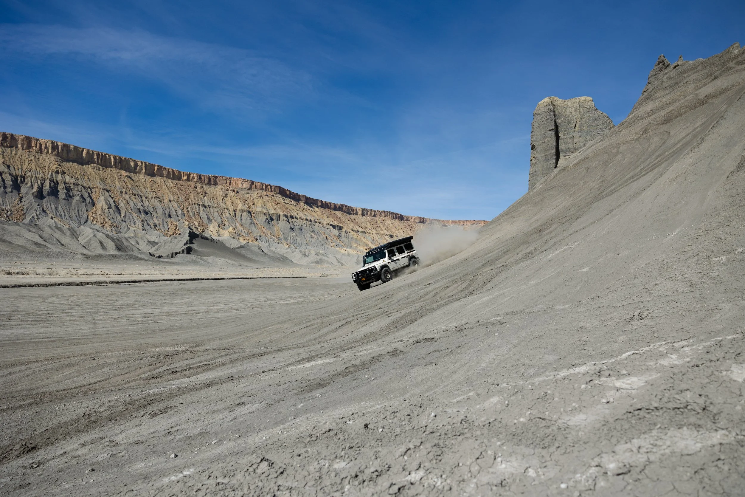 A rugged off-road vehicle driving up a steep sandy slope in a barren desert landscape with eroded rock formations in the background under a clear blue sky.