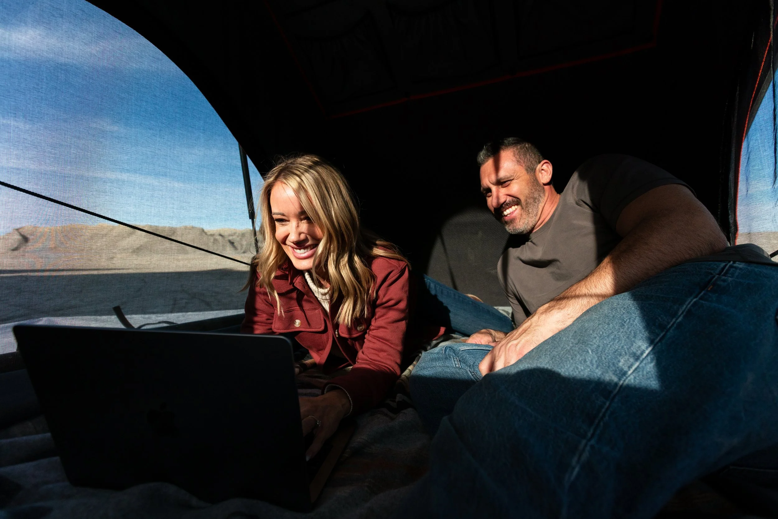 A man and woman are lying inside a camper, looking at a laptop and smiling. The woman has blonde hair and is wearing a rust-colored jacket. The man has short dark hair and is wearing a gray t-shirt. The camper has mesh windows with a view of a desert