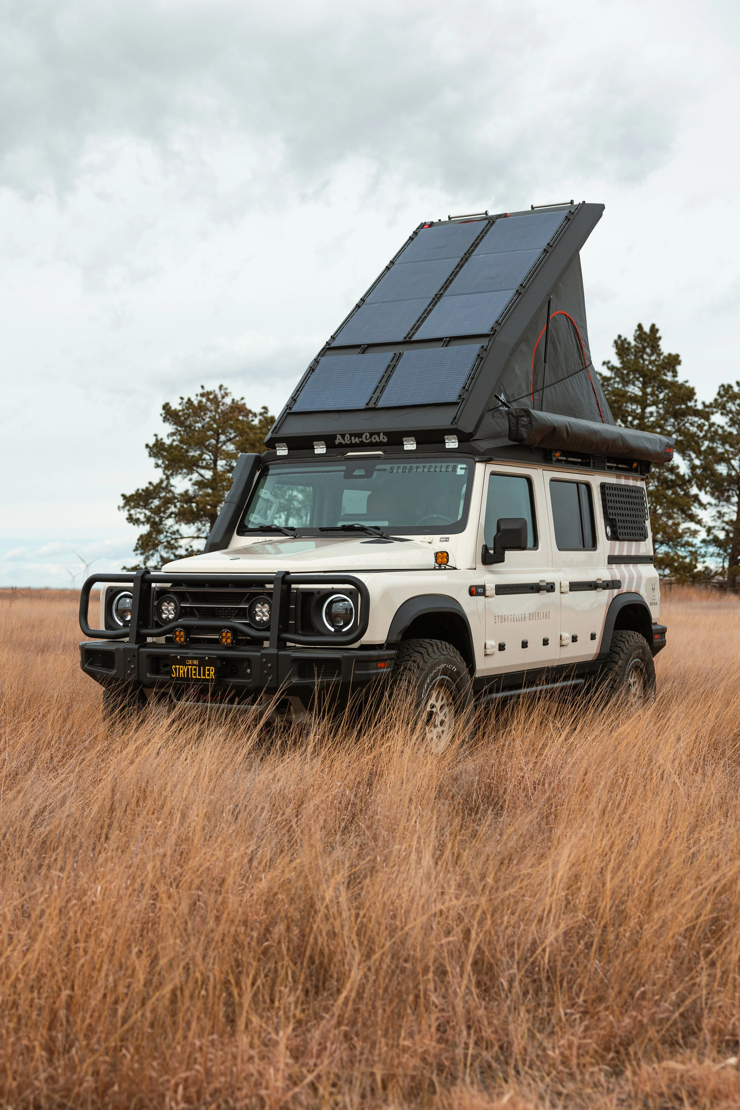 A white off-road vehicle with a roof-mounted tent and solar panels, parked in a grassy field with trees in the background.