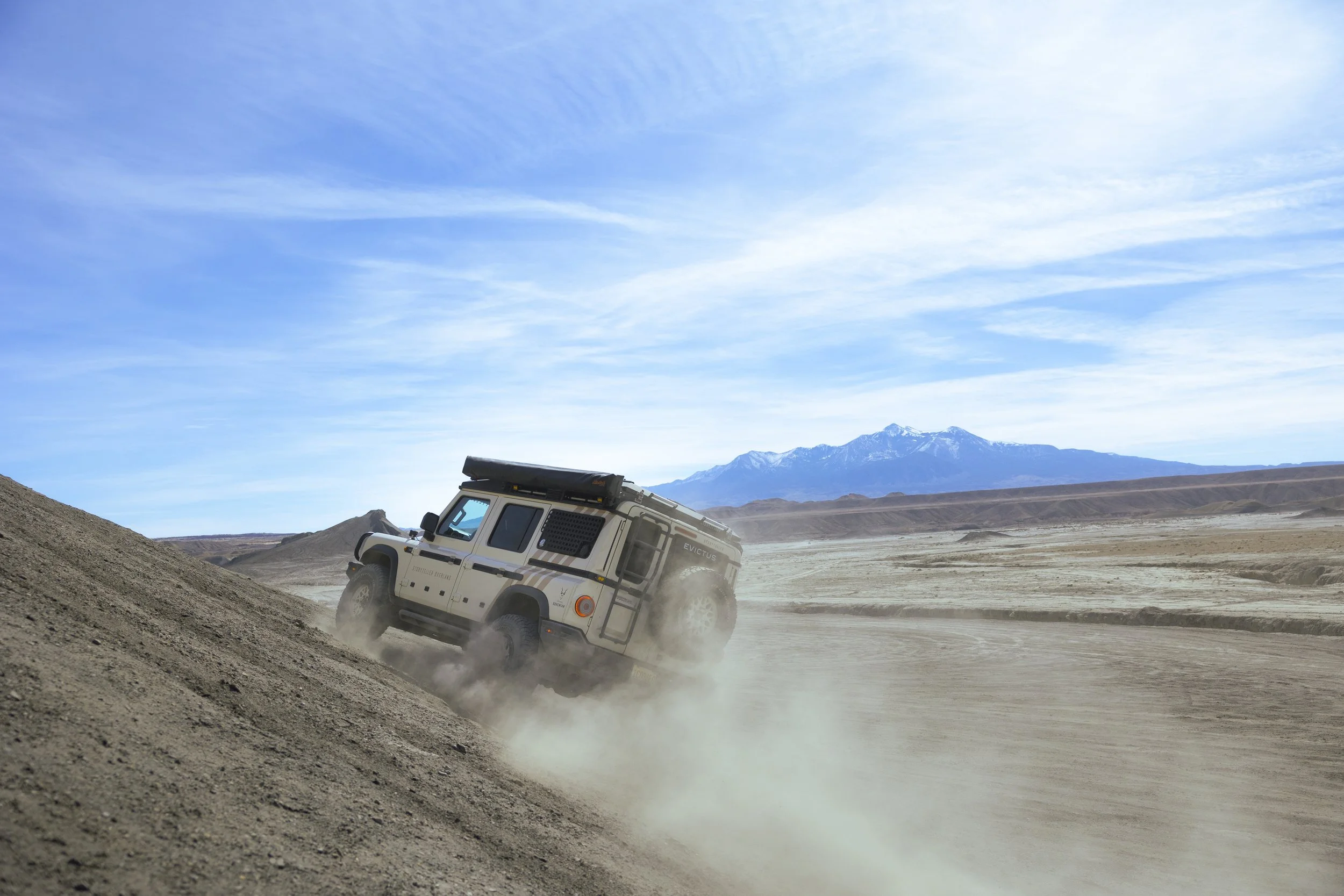 A white off-road vehicle climbing a dirt hill in a desert landscape under a blue sky with wispy clouds, mountains in the background.