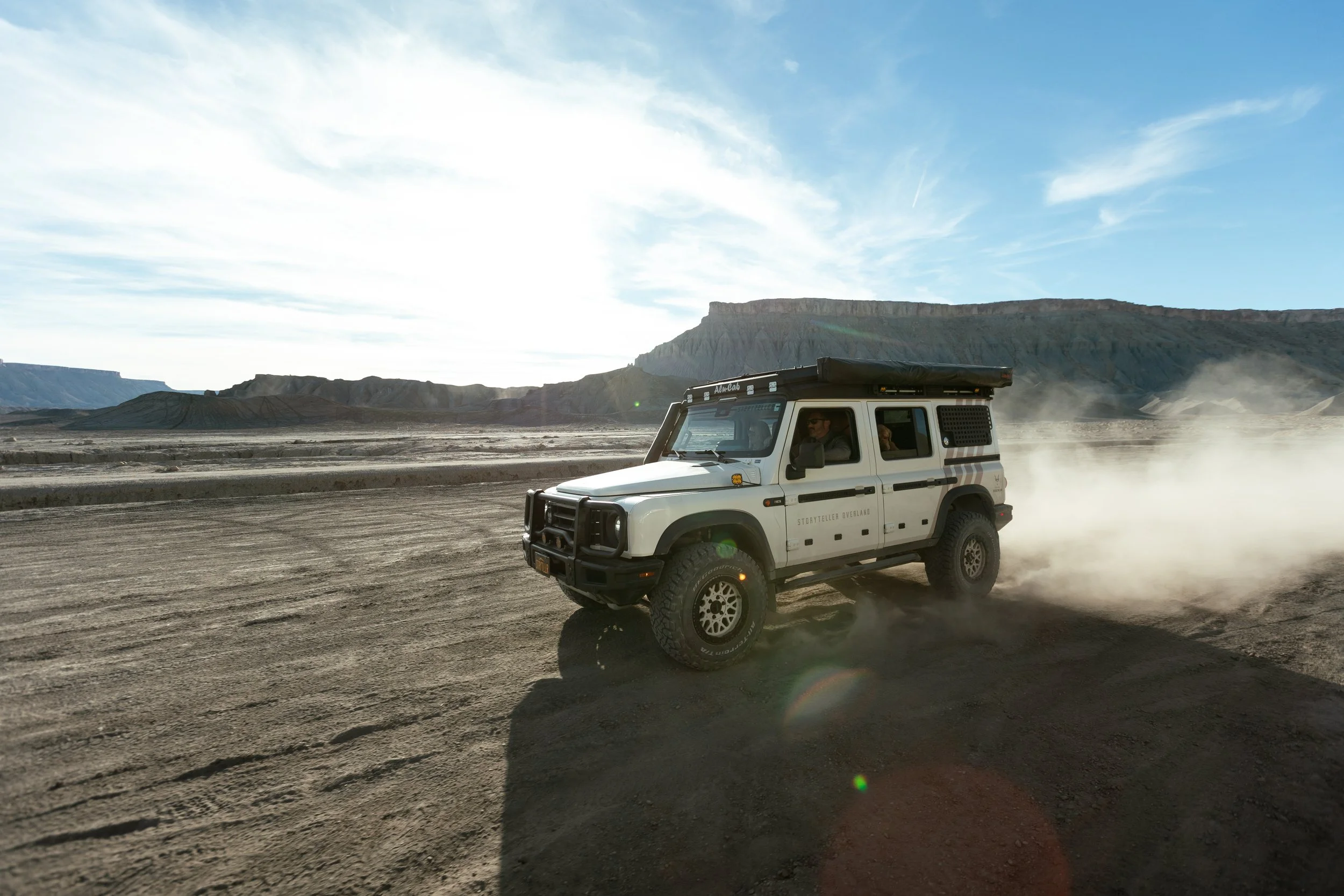 A white off-road vehicle driving on a dusty dirt road in a desert landscape with mountains in the background and a clear blue sky.