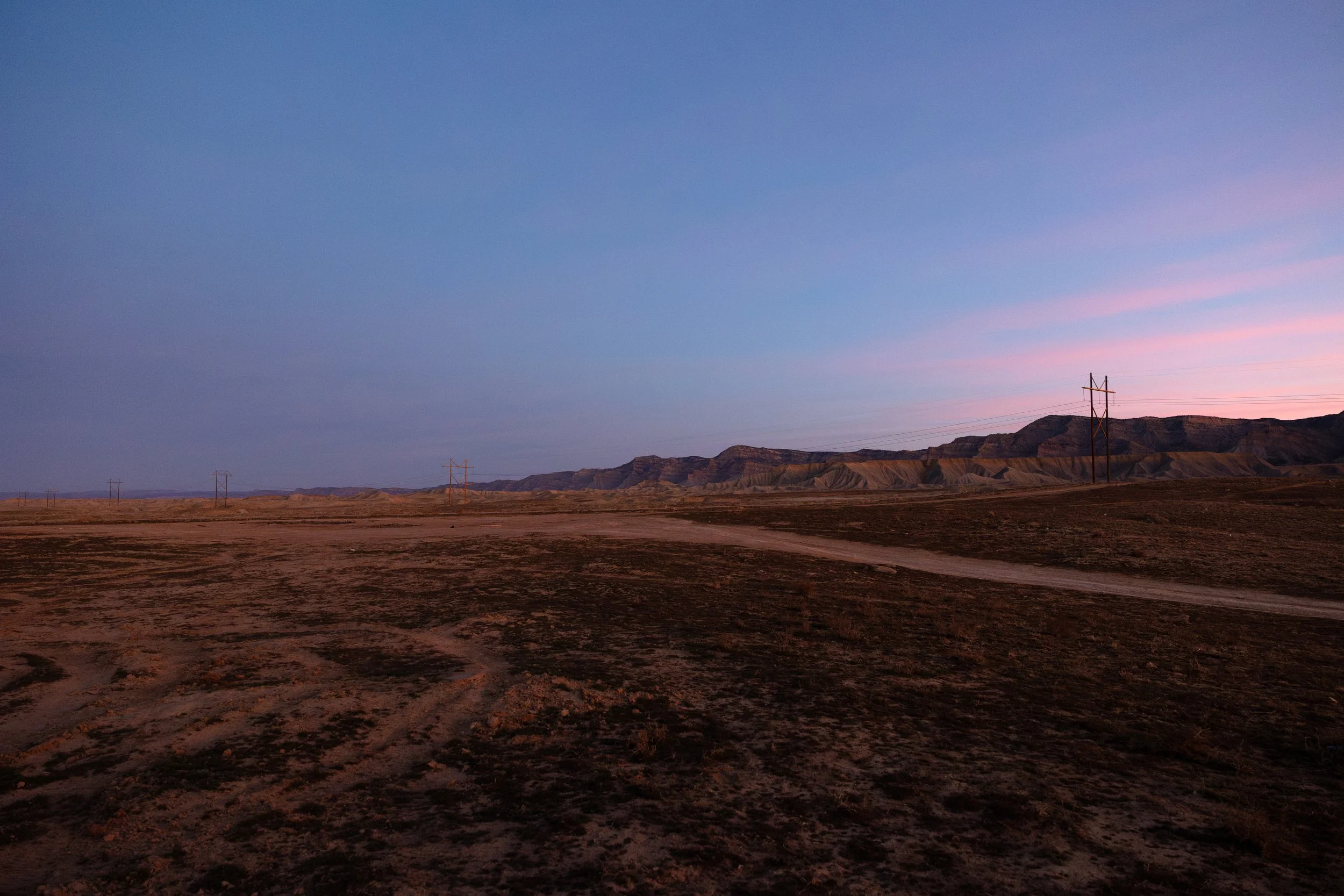 A vast, arid landscape with power lines extending into the distance. The terrain is flat with tracks, and there are distant mountains under a clear sky during sunset or sunrise.