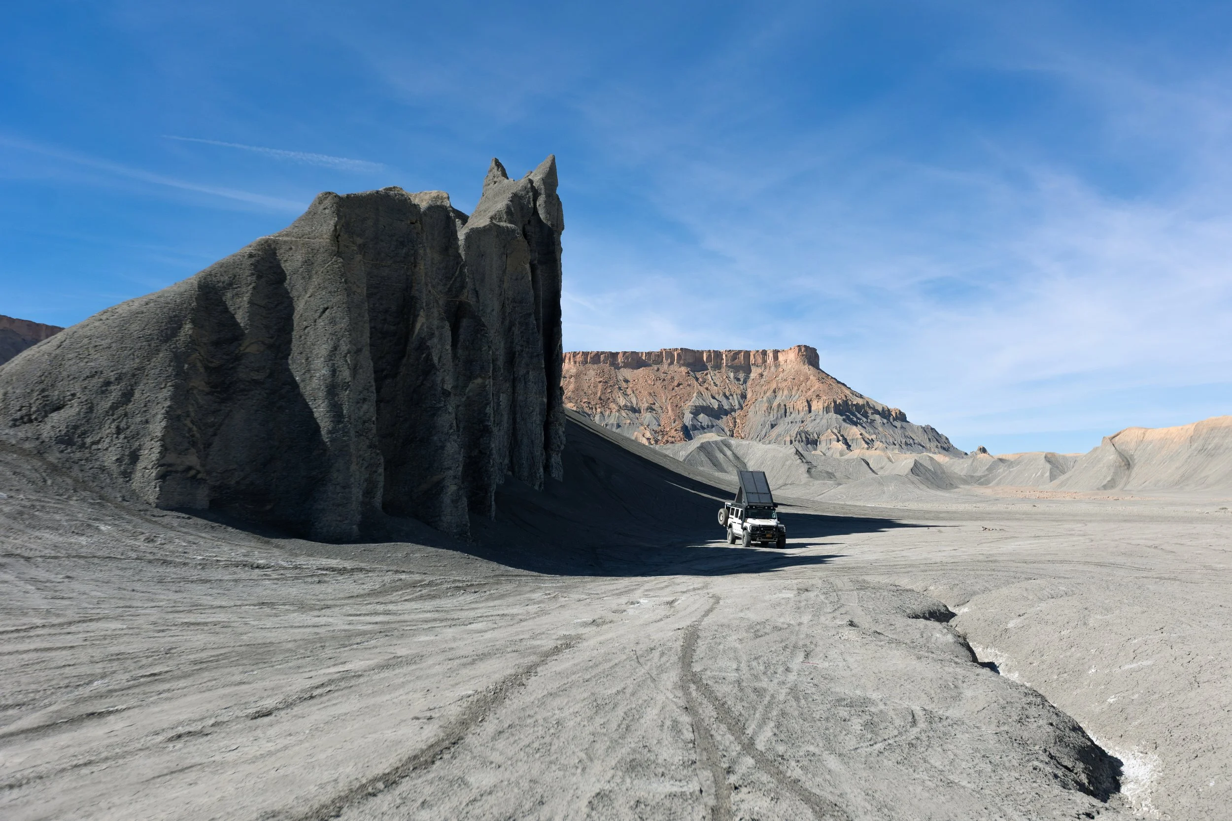 A rugged, barren desert landscape with large rock formations and a pickup truck with a solar panel set up on top, under a clear blue sky.