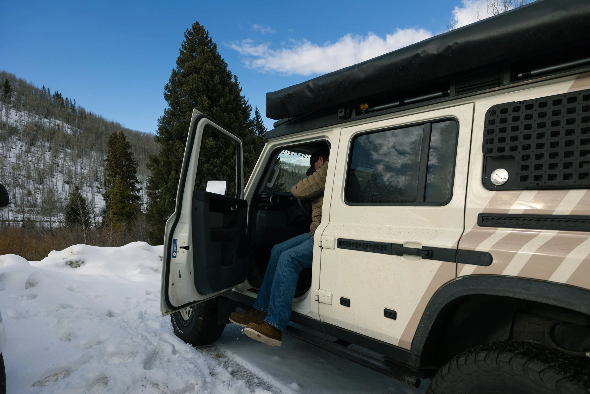 Person sitting inside the open door of a white off-road vehicle, parked on snow in a mountainous area with trees and blue sky in the background.