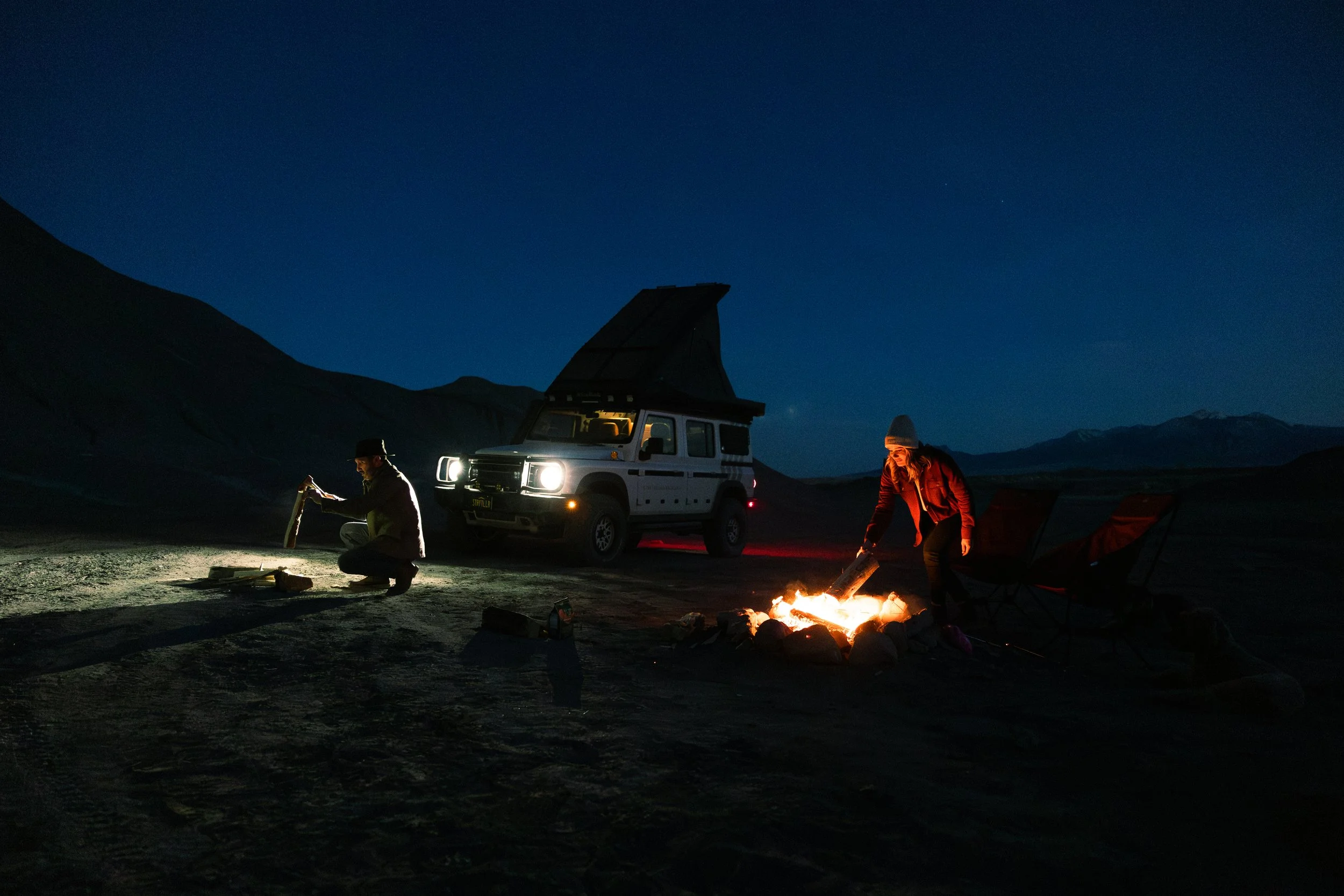 Two people camping at night near a vehicle with a rooftop tent, sitting by a campfire in a mountainous area.