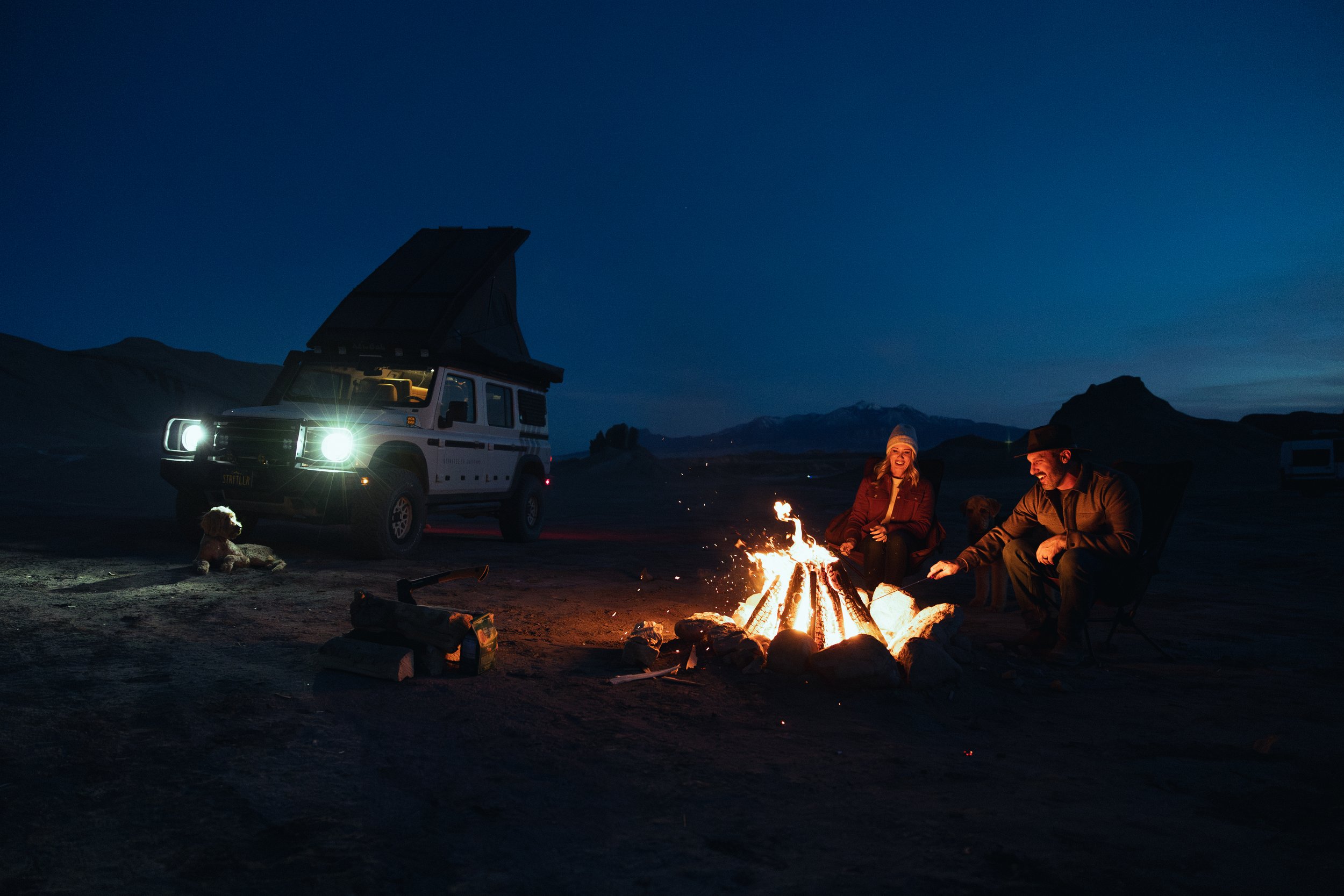 Two people sitting around a campfire at night with a dog and a parked camper van in the background.
