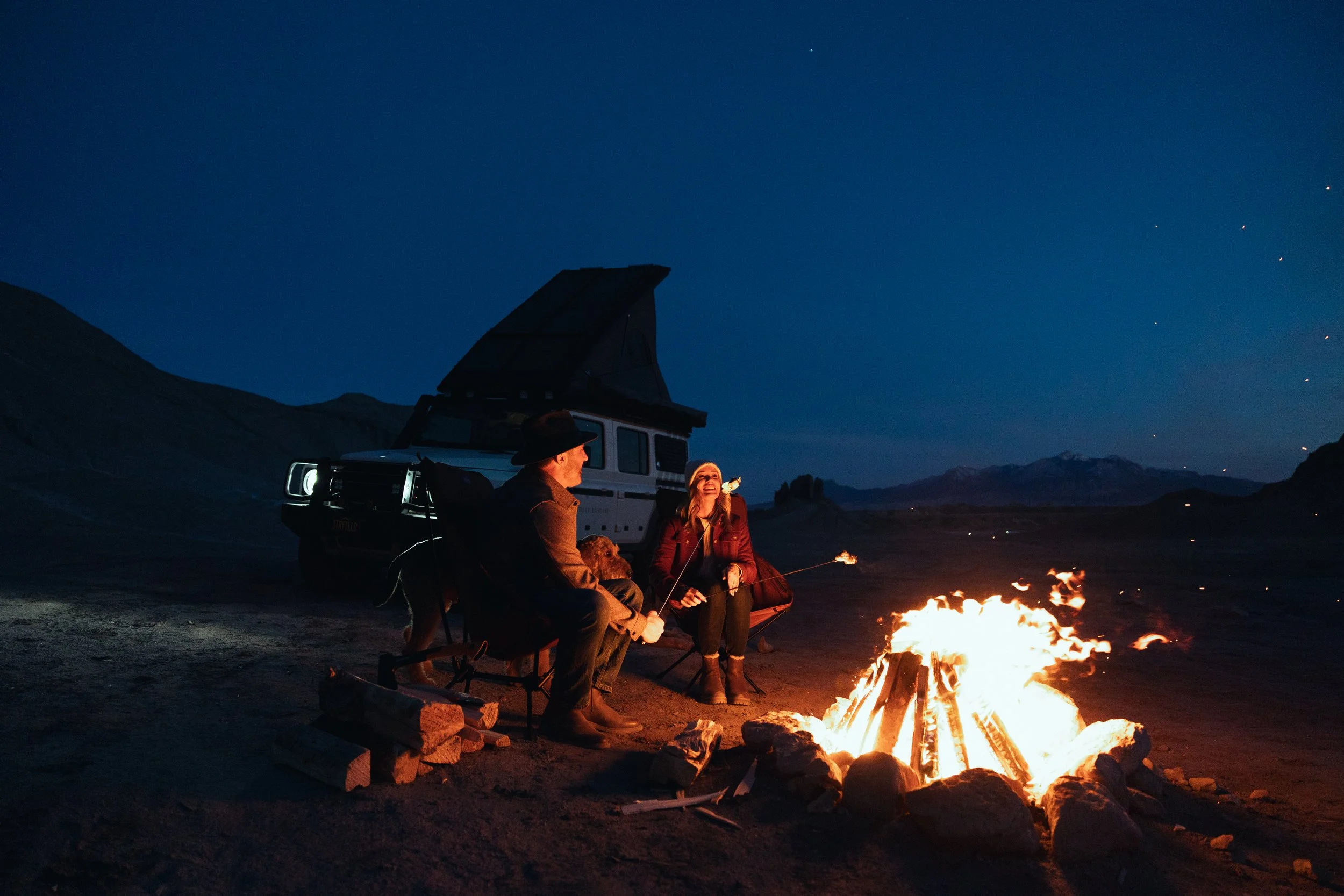 Two people sitting by a campfire in a desert at night with a camper trailer nearby, mountains in the background, starry sky overhead.