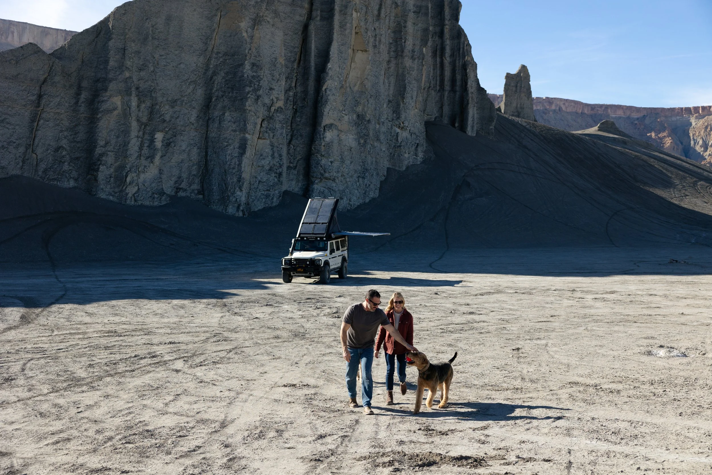 Two people and a dog walking on desert terrain with a vehicle and large rocky cliffs in the background.