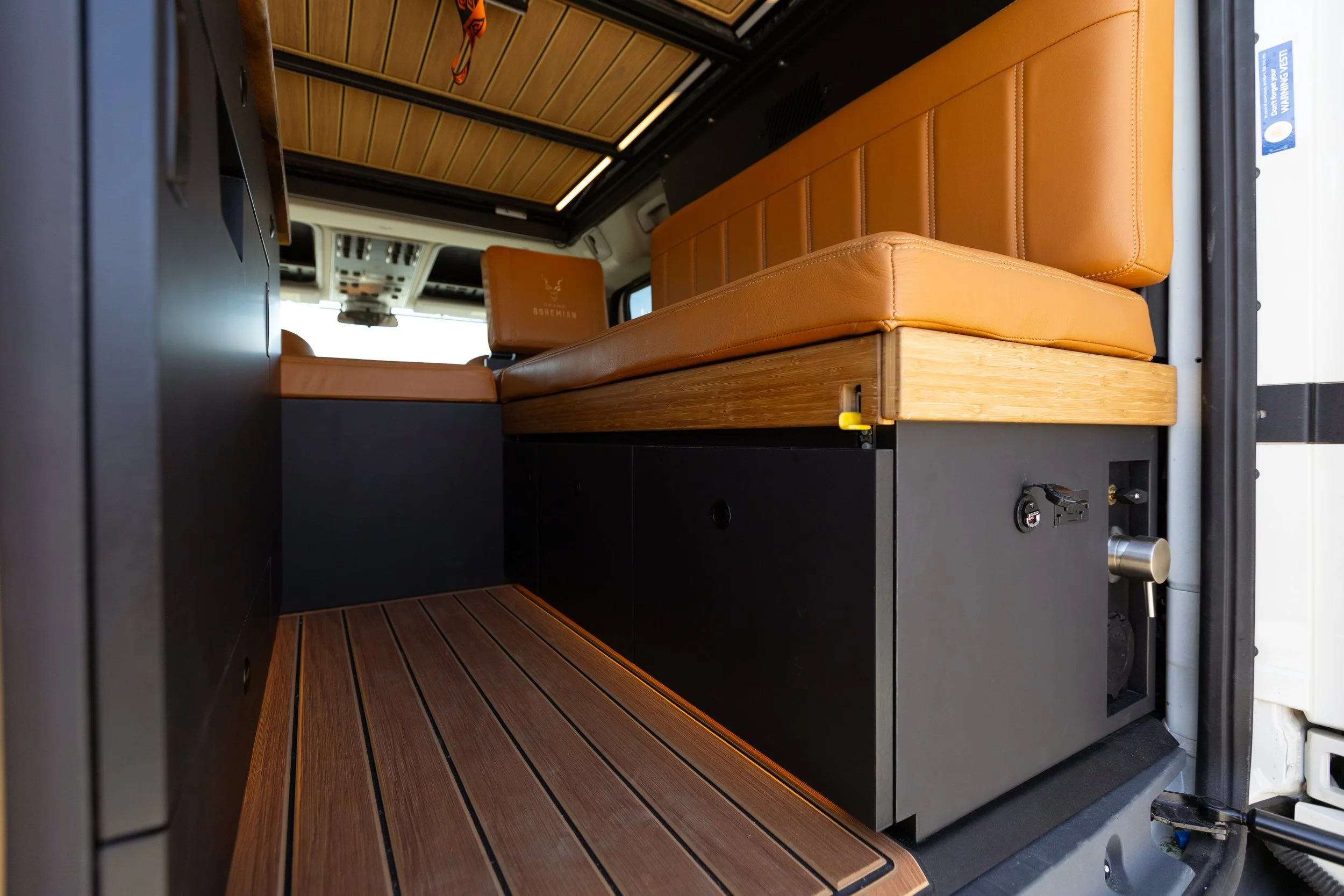 Interior of a van with a custom wooden floor and tan leather cushioned seats. The ceiling has wooden paneling and there is a view of the driver and passenger seats in the front. The space looks designed for comfort and style.