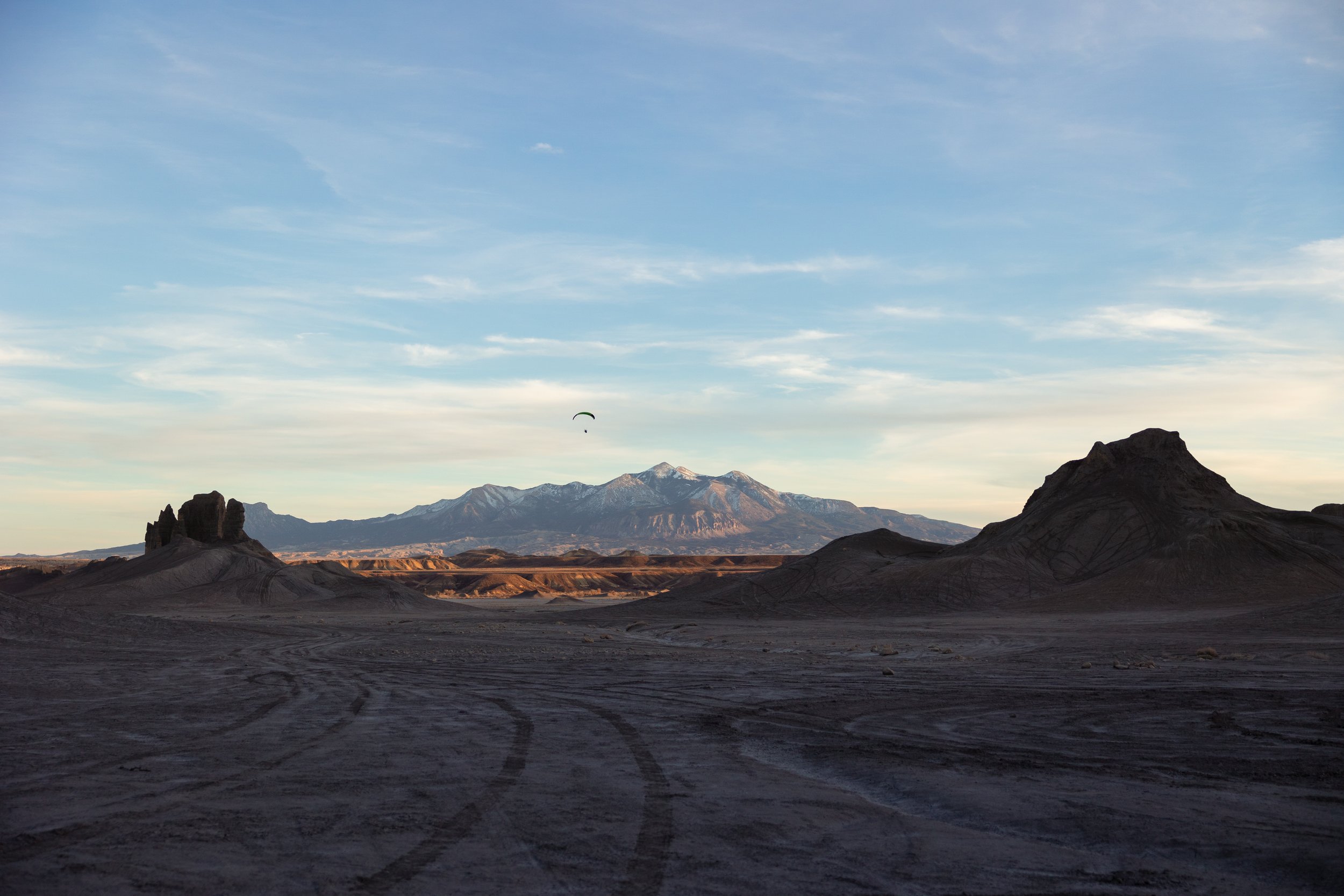 Desert landscape with rugged hills and mountains in the background, a sky with wispy clouds, and a paraglider flying above.