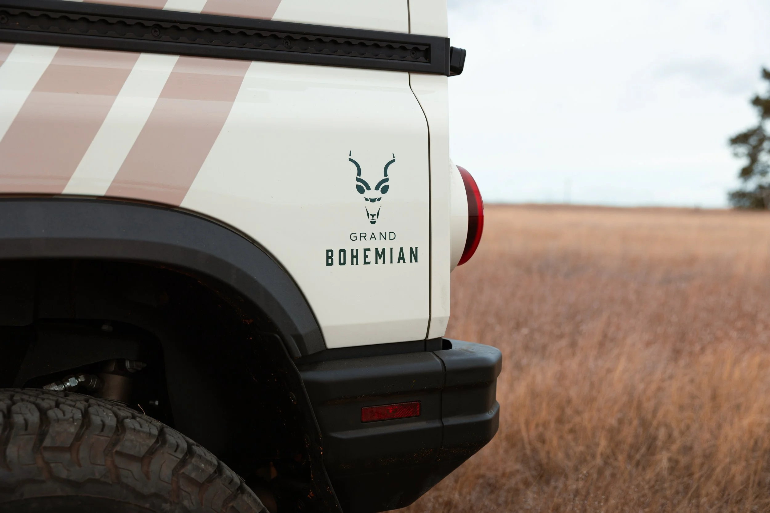 Close-up of the rear side of a Jeep with a Bohemian-themed decal, showing part of the rear wheel, taillight, and a background of an open field and cloudy sky.