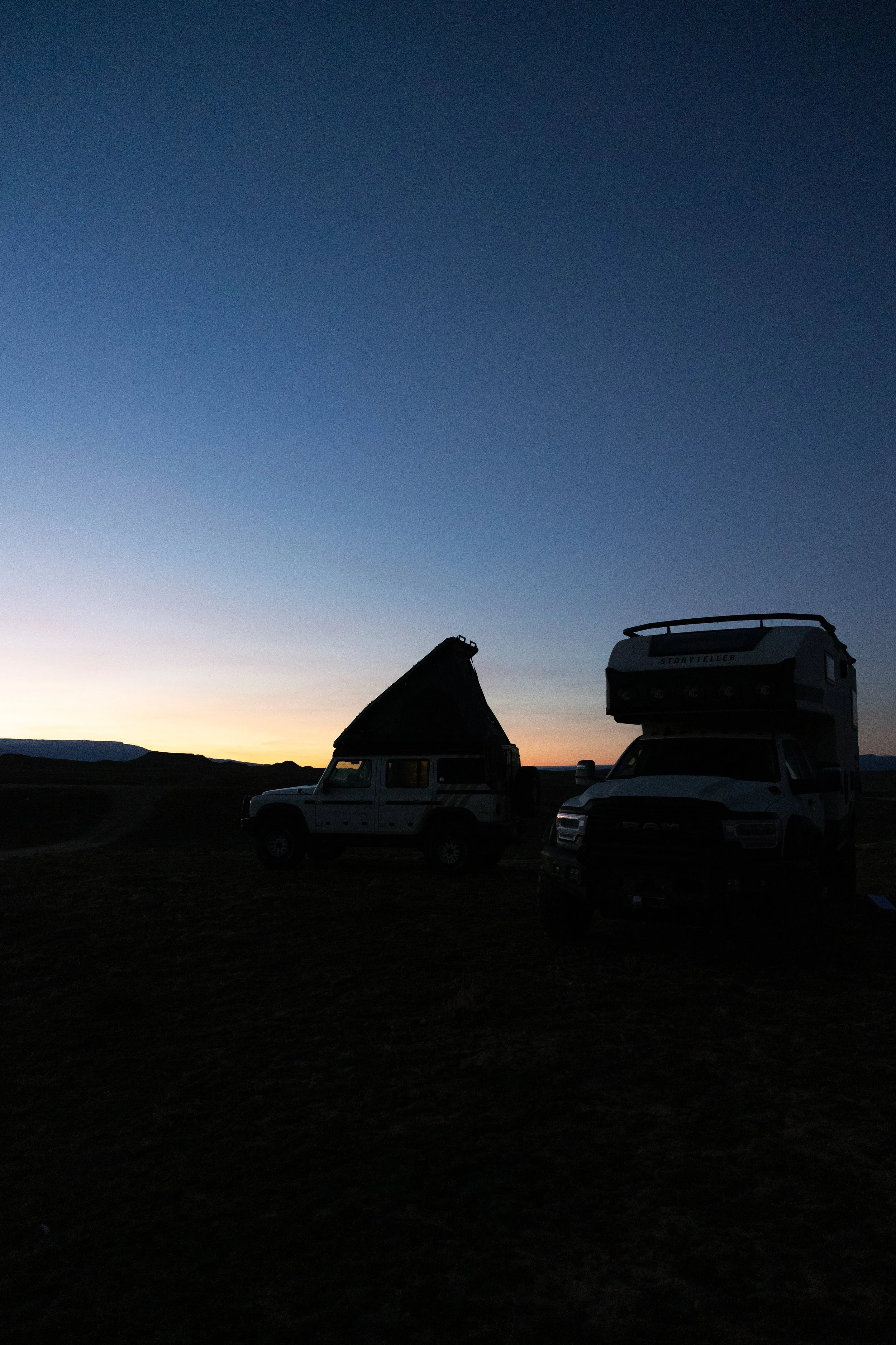 Silhouettes of two off-road vehicles with a camper on a mountain landscape during dawn or dusk, with a gradient sky from orange to deep blue.