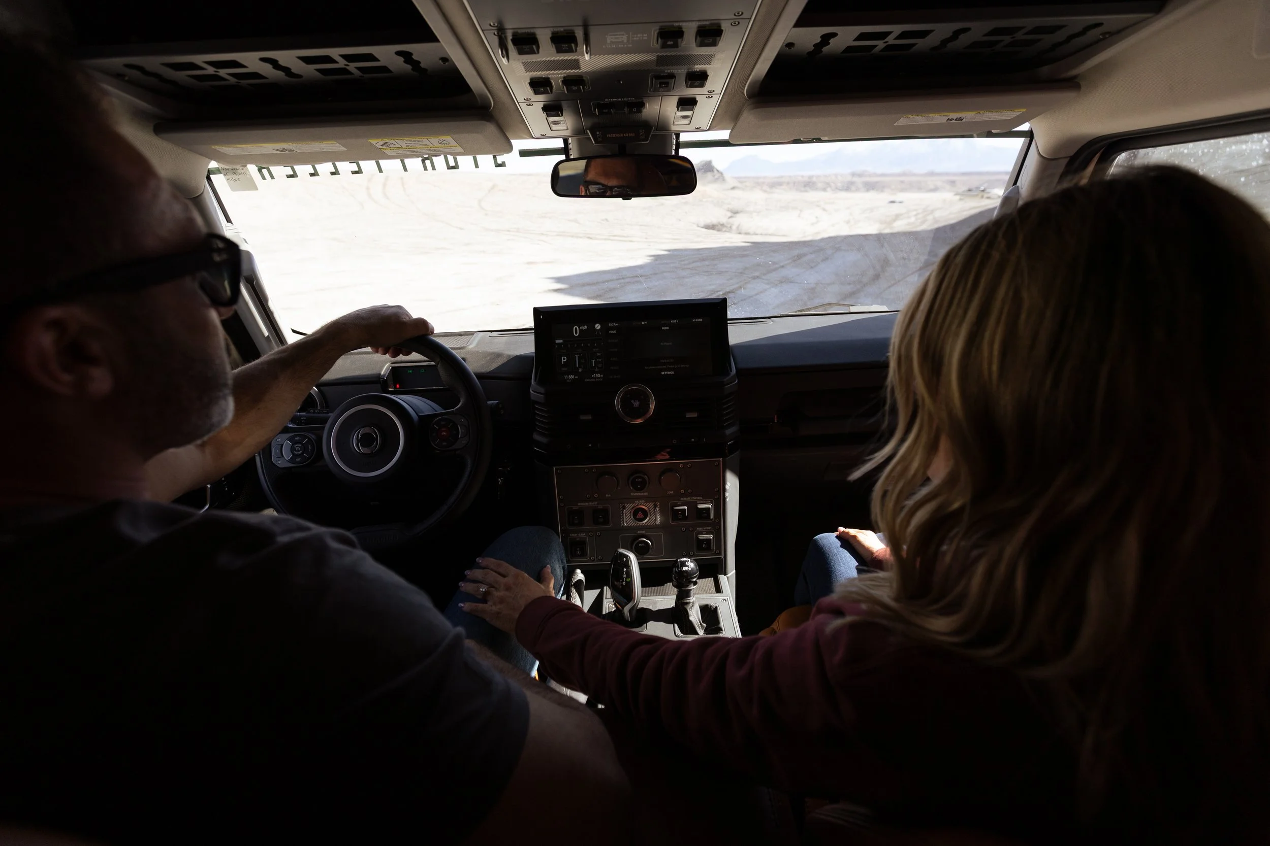 Inside view of a vehicle with two passengers, one driving and the other a passenger, on a dirt road in a desert landscape.