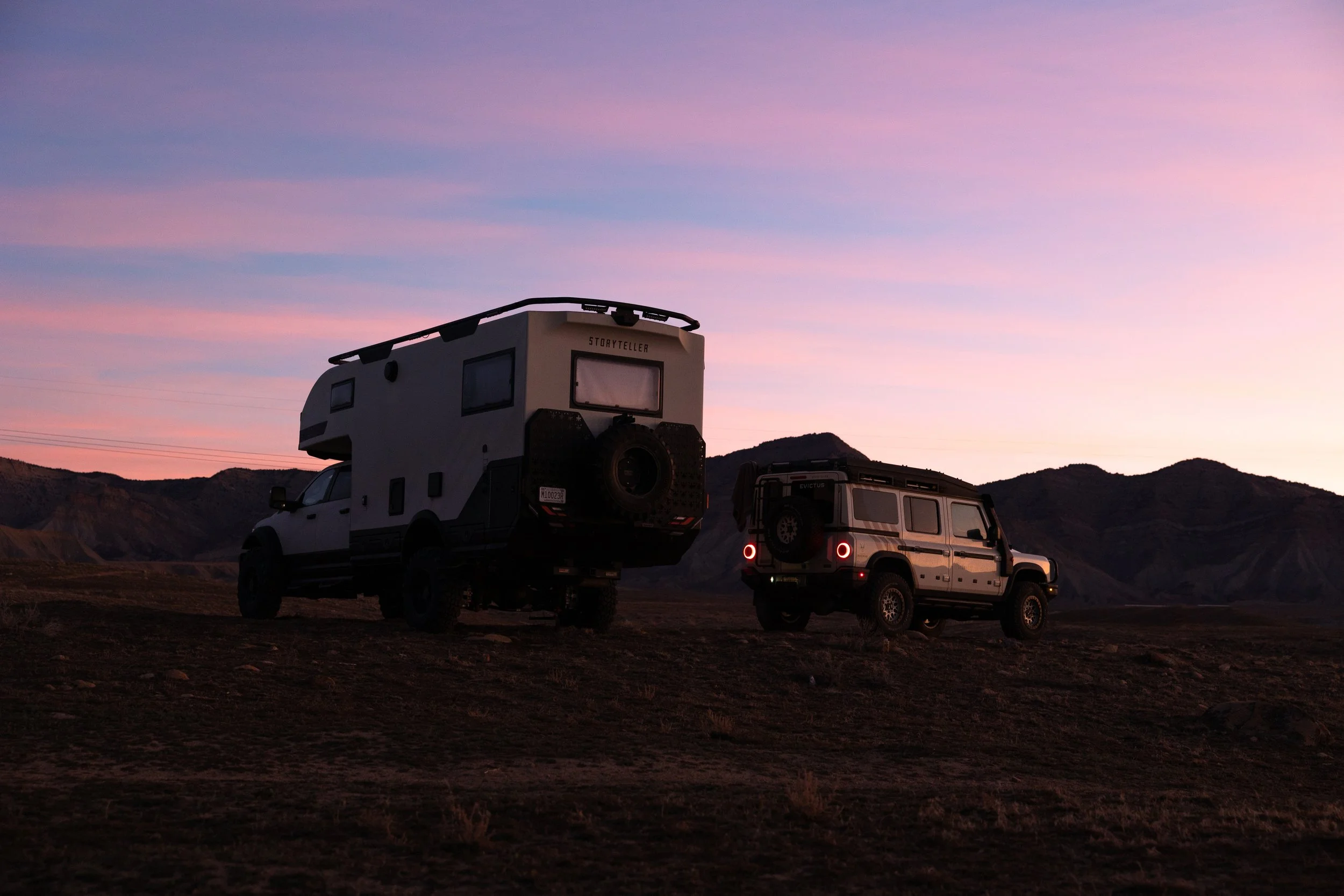 Two off-road vehicles parked on a rocky landscape during sunset, with mountains in the background and a sky with pink and purple hues.