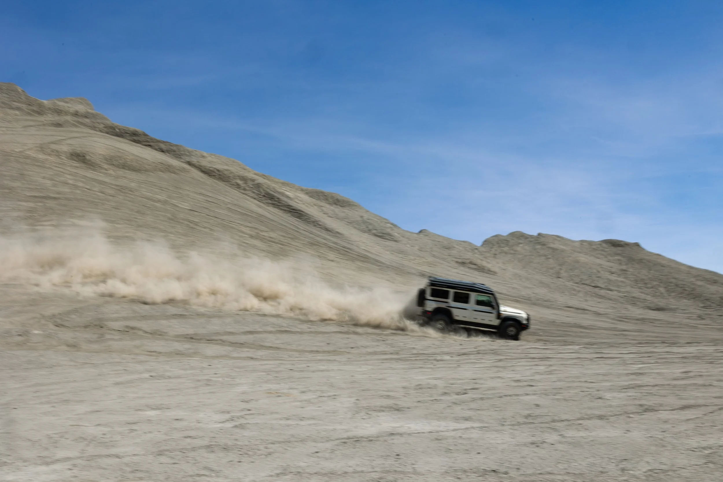 A white off-road vehicle driving across a desert landscape, kicking up dust, under a blue sky.
