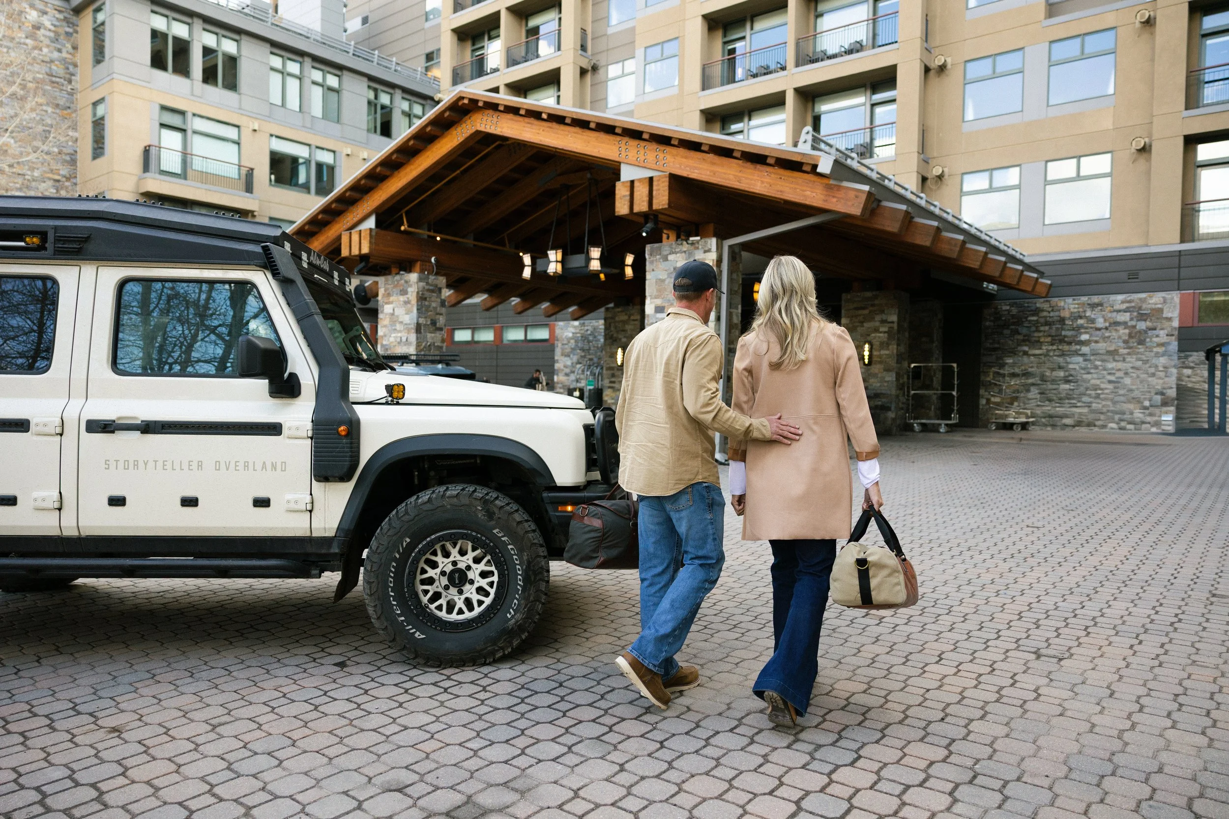A man and a woman walking towards an entrance of a building with a covered wooden roof, with a white off-road vehicle parked nearby. The man has a black cap, beige jacket, and jeans, and the woman has long blonde hair, a beige coat, and is carrying a