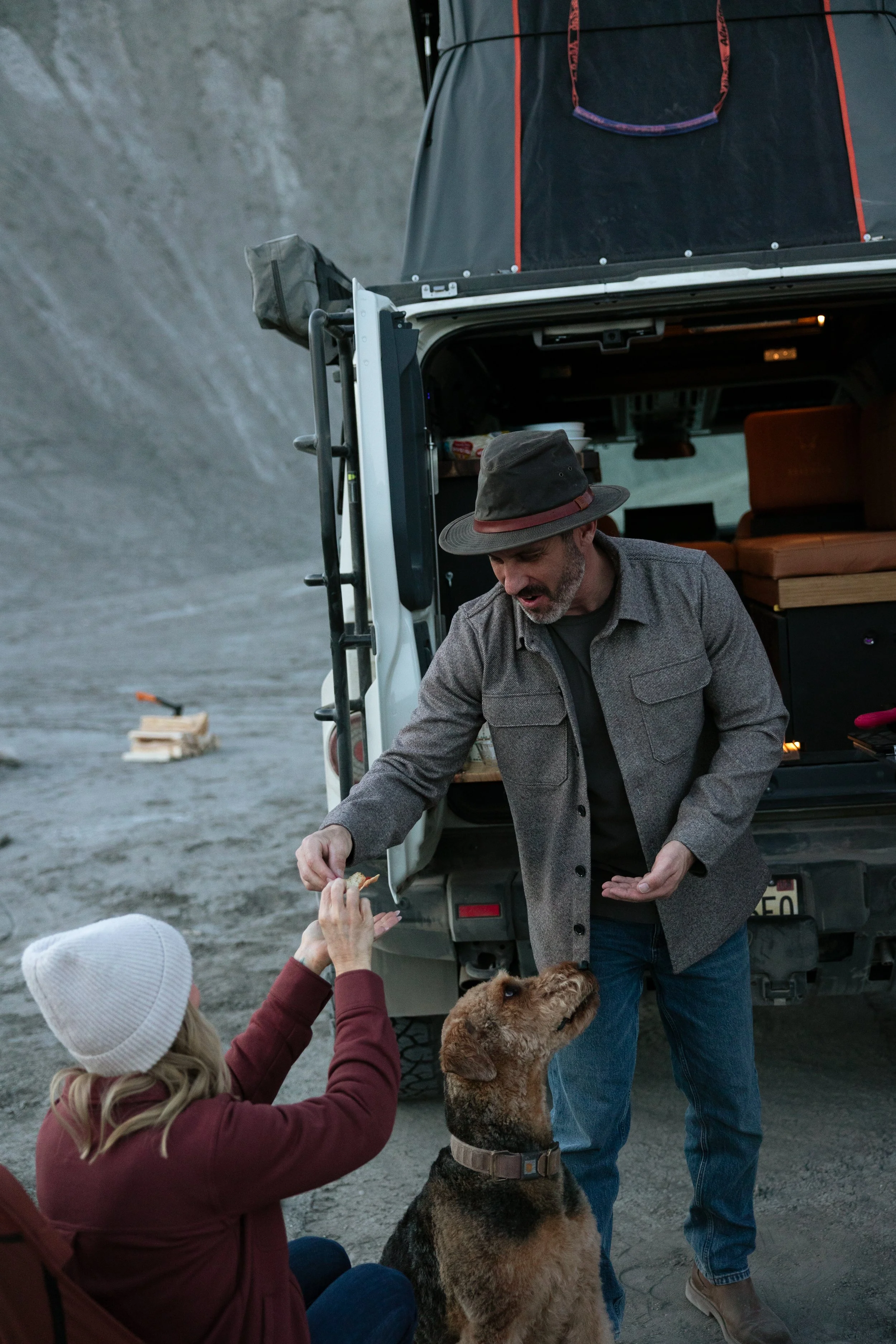A man with a beard and a woman wearing a white knit hat are outdoors near an off-road vehicle with a rooftop tent. The woman is giving a dog a treat, and the dog, a large breed, is sitting attentively.