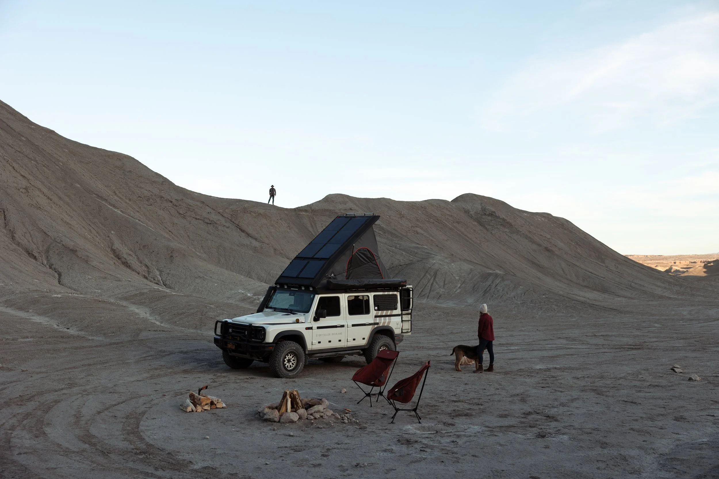 A person and a dog near a white off-road vehicle with solar panels on top, parked in a desert landscape with barren hills and firewood arranged in a circle nearby. Another person stands on a hill in the distance.