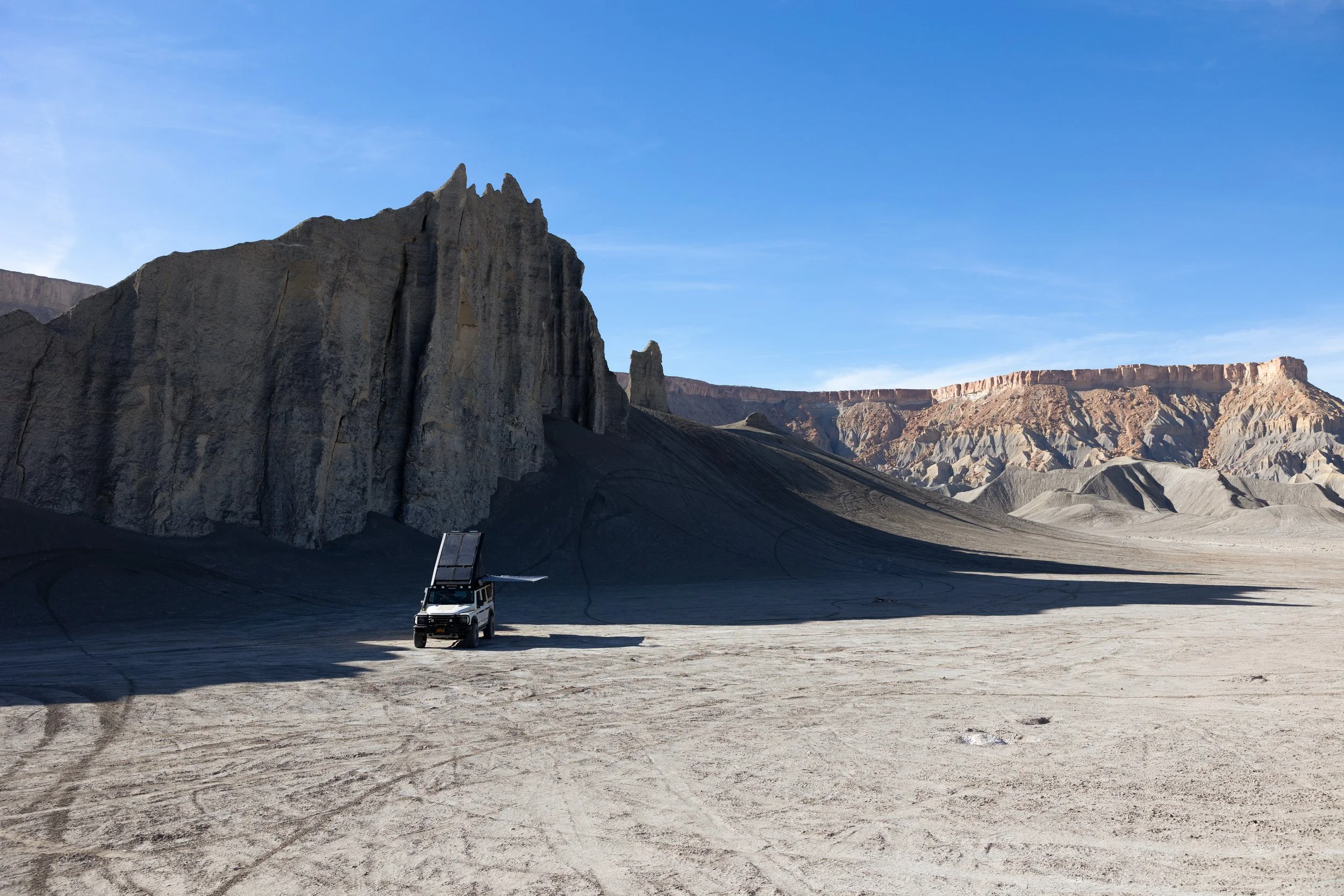 A rugged desert landscape with rocky cliffs and mesas under a blue sky. A black off-road vehicle with solar panels mounted on top is parked on the flat, sandy terrain.