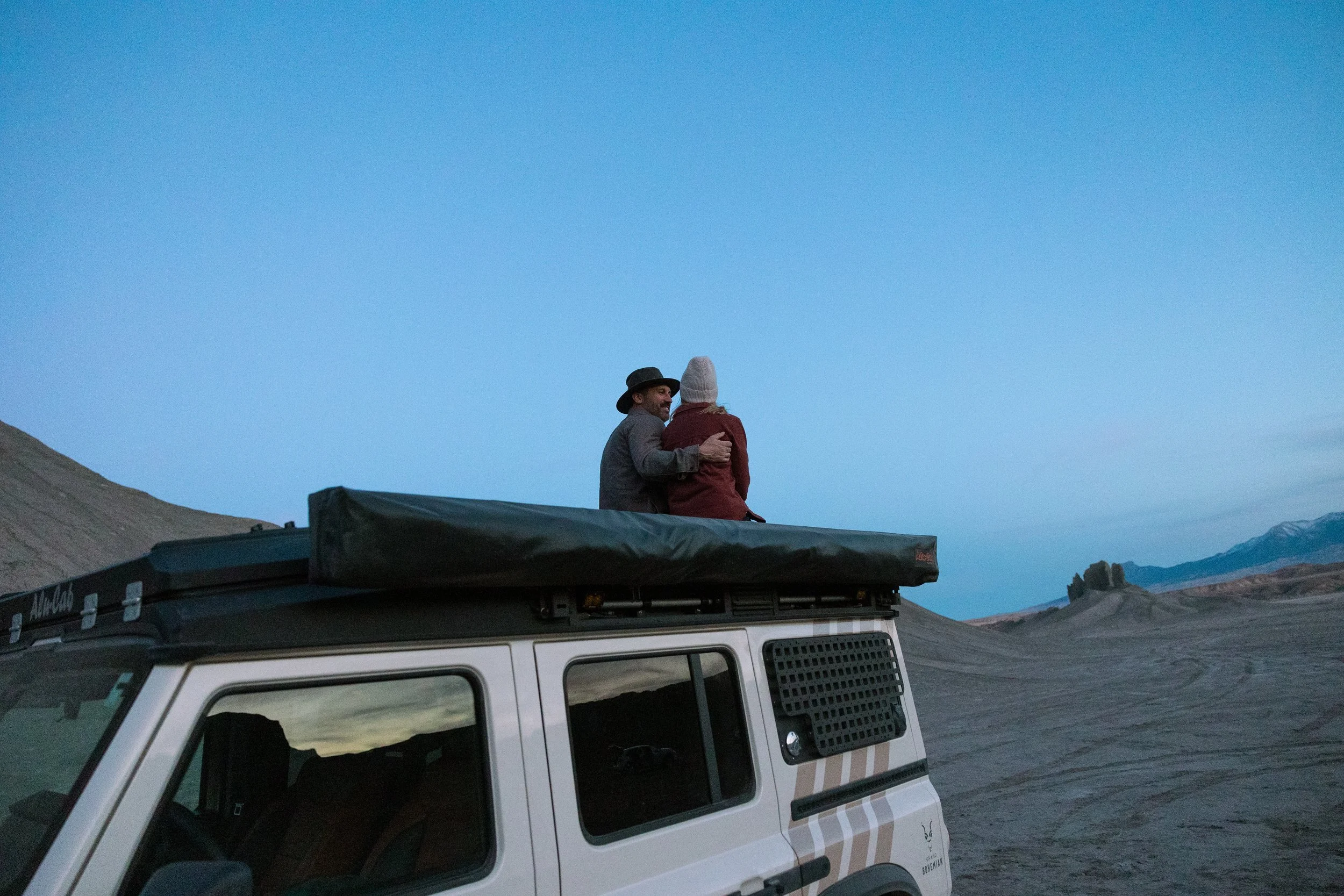 A man and a woman sitting on top of a vehicle in a desert landscape at dusk, with mountains in the background.