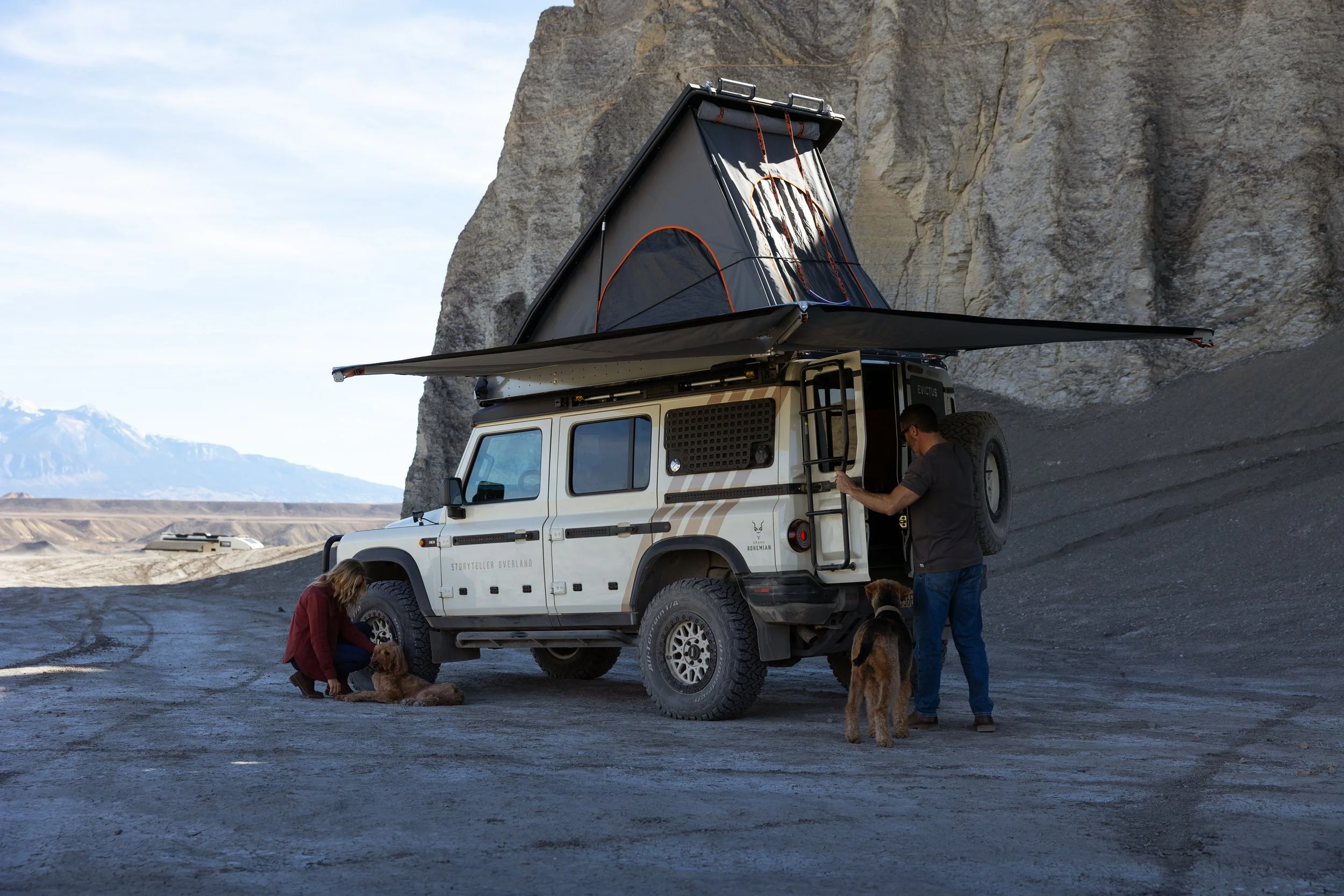 People with dogs next to a white off-road vehicle with a rooftop tent installed, parked near a rocky cliff in a desert landscape.