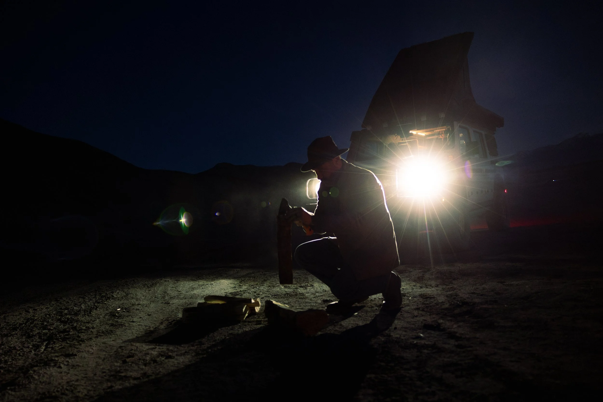 A silhouette of a person wearing a hat, kneeling on the ground and working with tools under a bright vehicle headlights at night in a mountainous area.