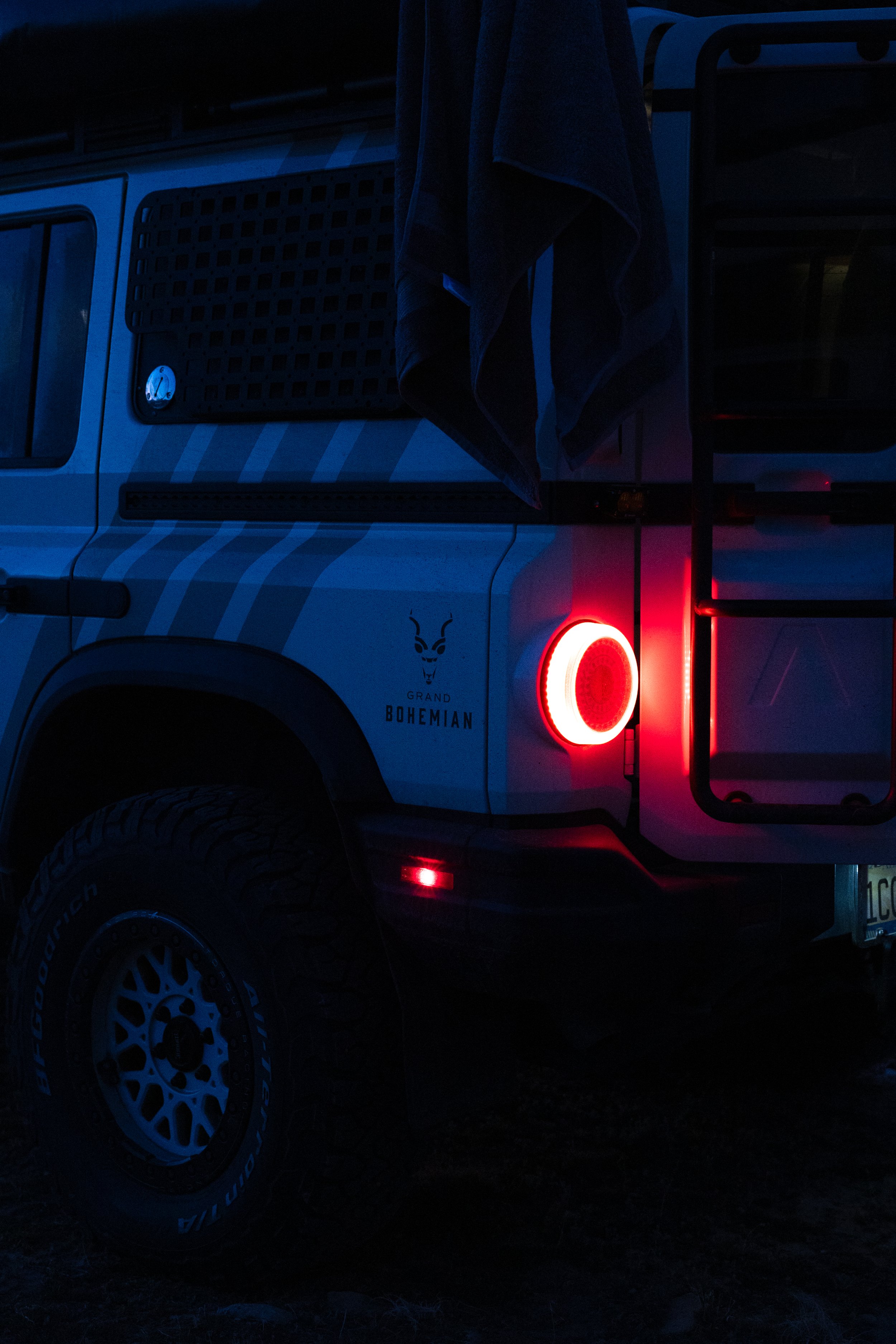 The rear of a white off-road vehicle with a large tire, a red taillight, and a decal that says 'Grand Bohemian' with a goat logo.