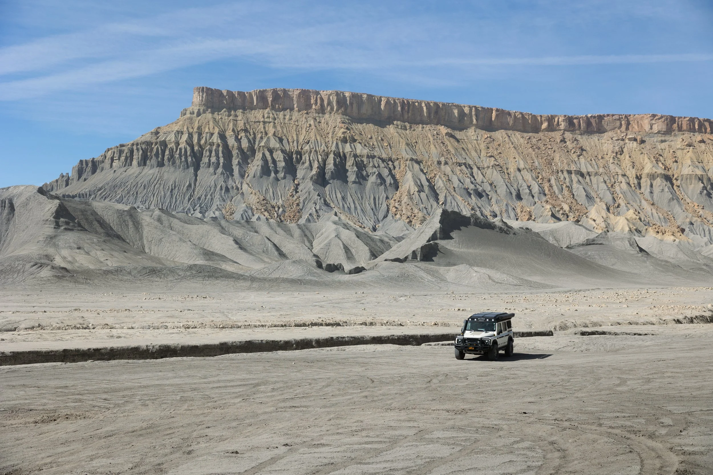 A white off-road vehicle driving on a sandy desert landscape with layered mountains in the background under a blue sky.