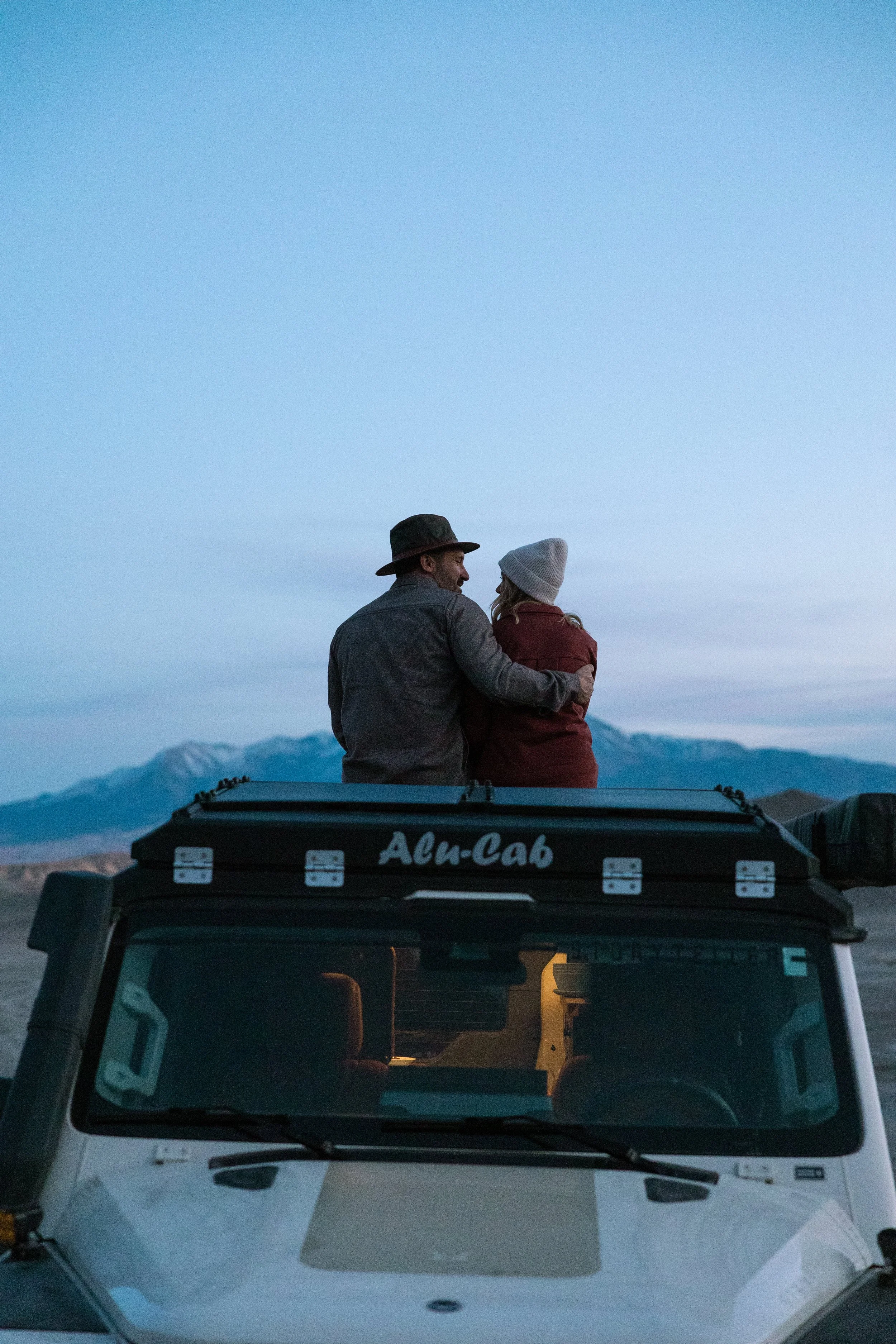 A couple sits on top of a white off-road vehicle labeled 'Alu-Cab' in a scenic mountainous landscape during dusk, with a mountain range visible in the background.