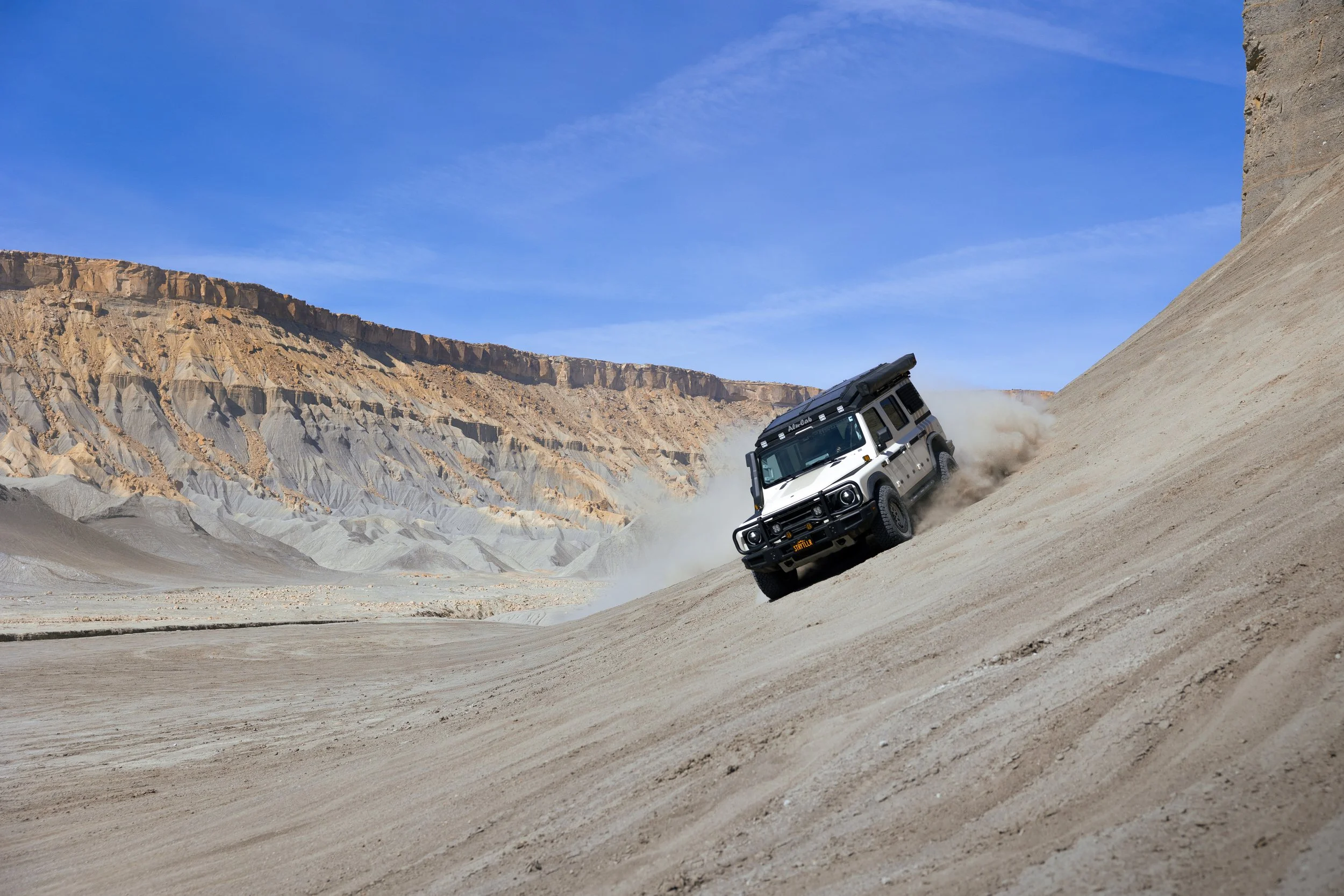 A white off-road vehicle driving up a steep sandy slope in a desert landscape with rugged cliffs and a blue sky in the background.