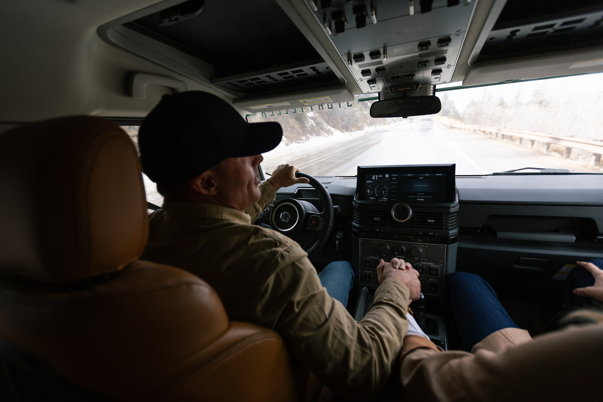 Inside a vehicle, a person wearing a hat and beige jacket drives on snowy roads, with a dashboard and a touchscreen display visible.