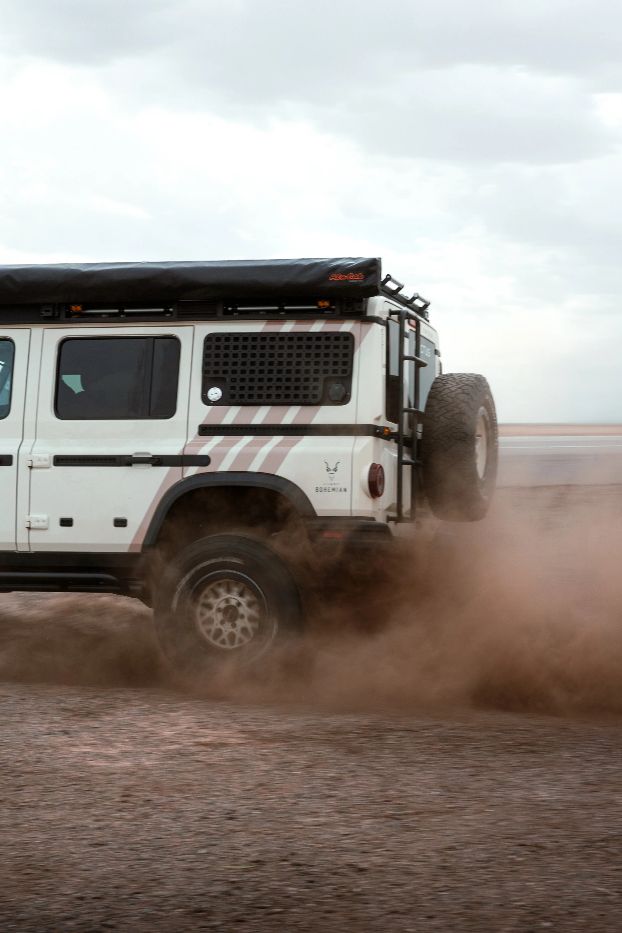 A white off-road vehicle driving on a dirt road, kicking up dust, with a spare tire mounted on the back.