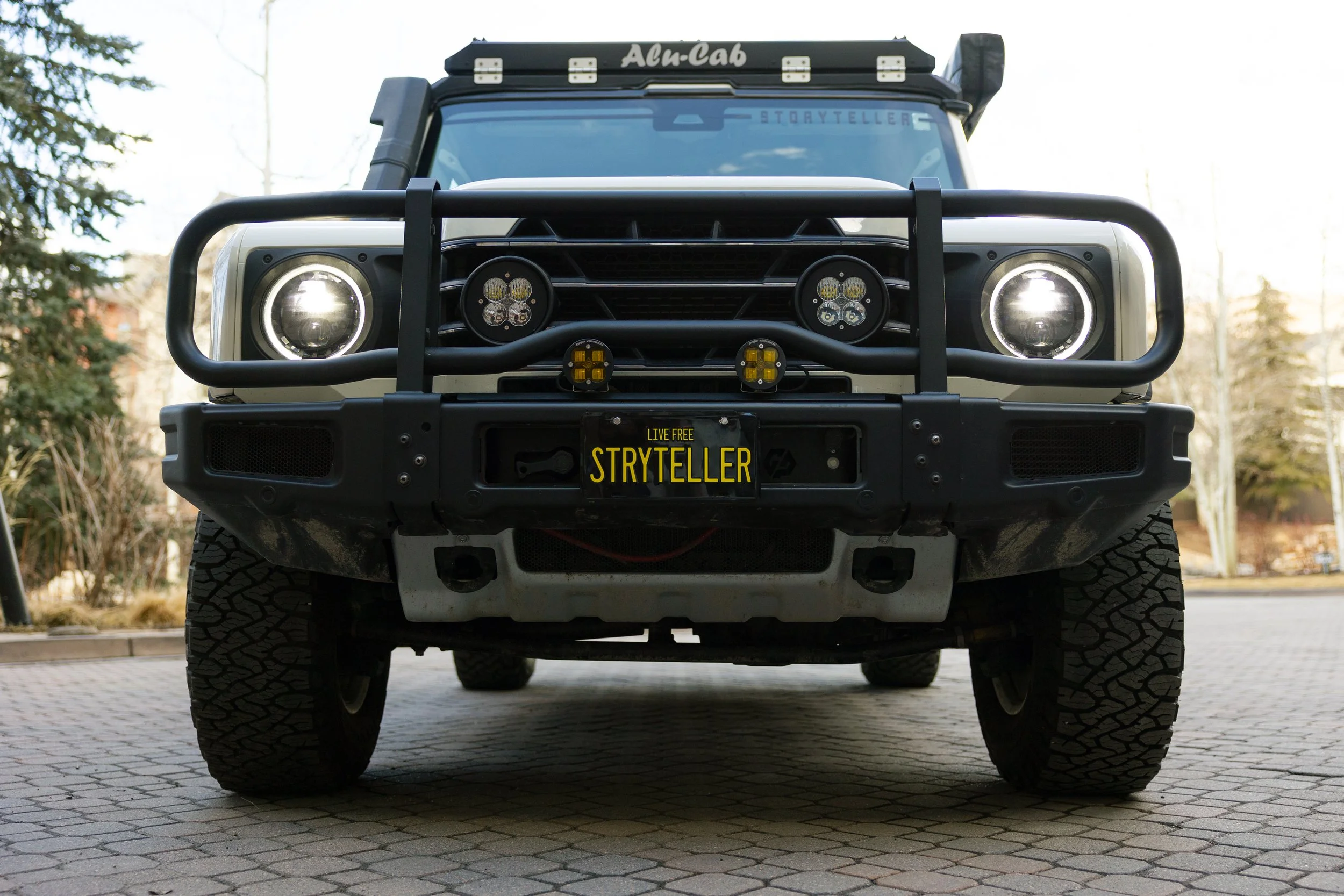 Front view of a black off-road vehicle with large tires, winch, and front bumper guard, parked on a paved street in a suburban area.