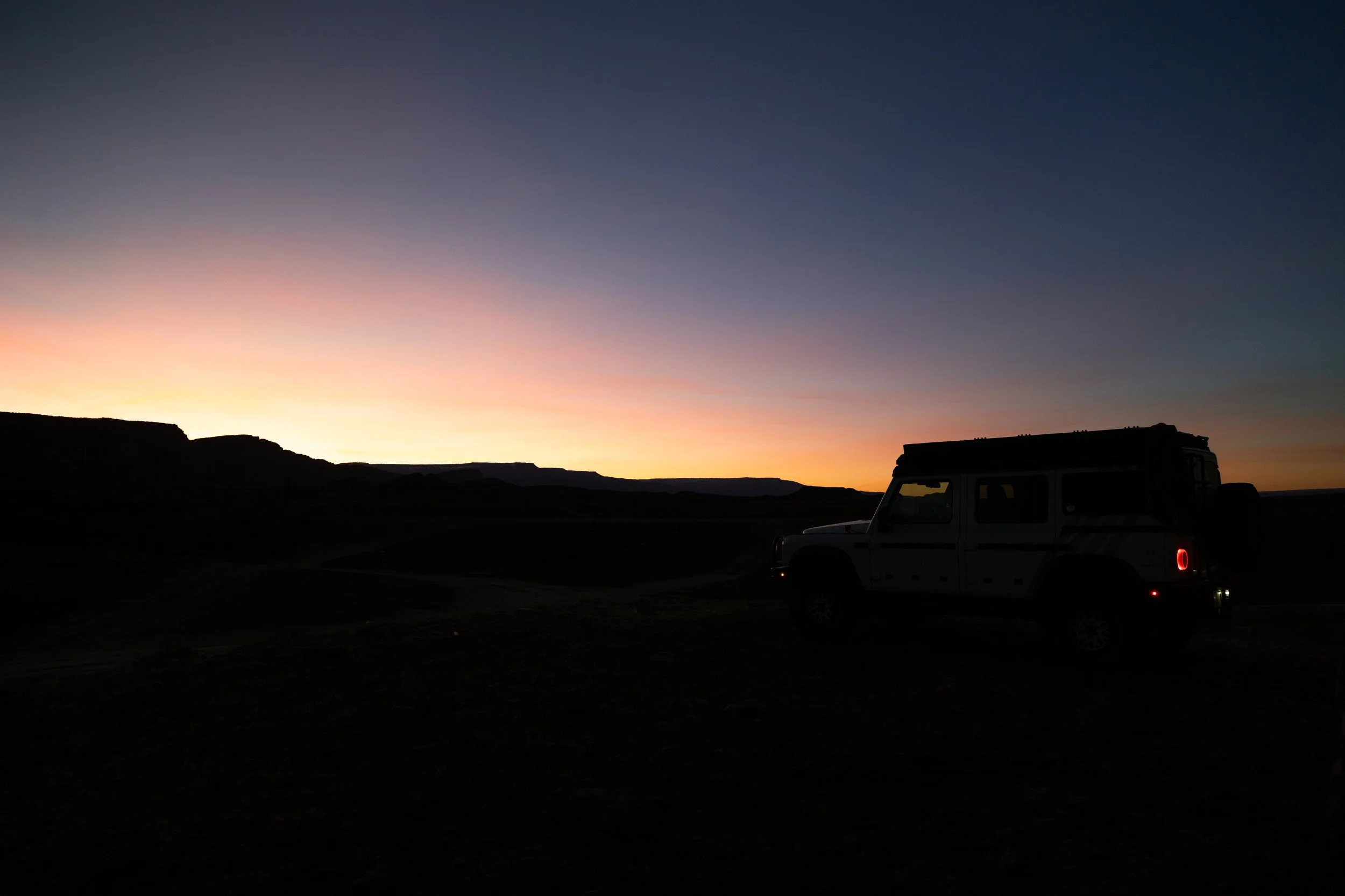 Silhouette of an off-road vehicle parked on a dark landscape at sunset, with a colorful sky and distant mountains in the background.