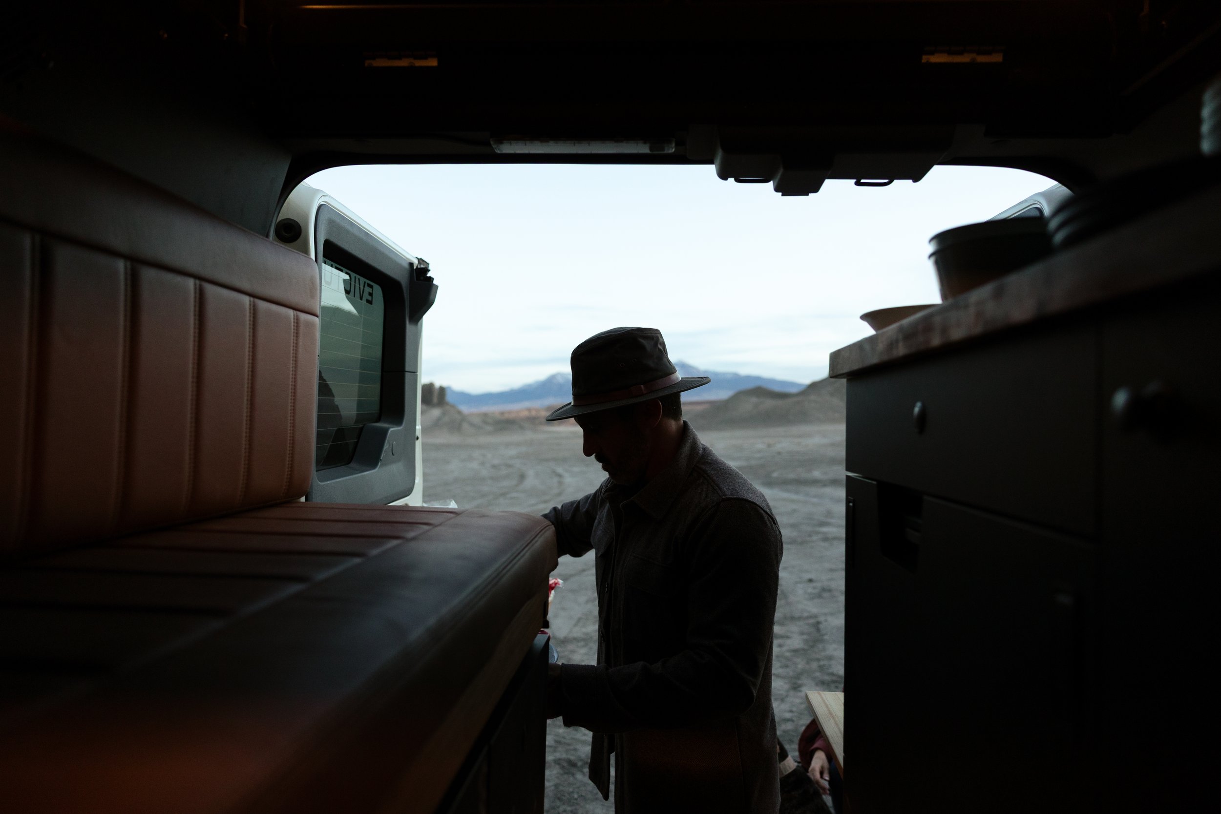 Silhouette of a man wearing a hat inside a vehicle, looking down, with a desert landscape and mountains in the background.